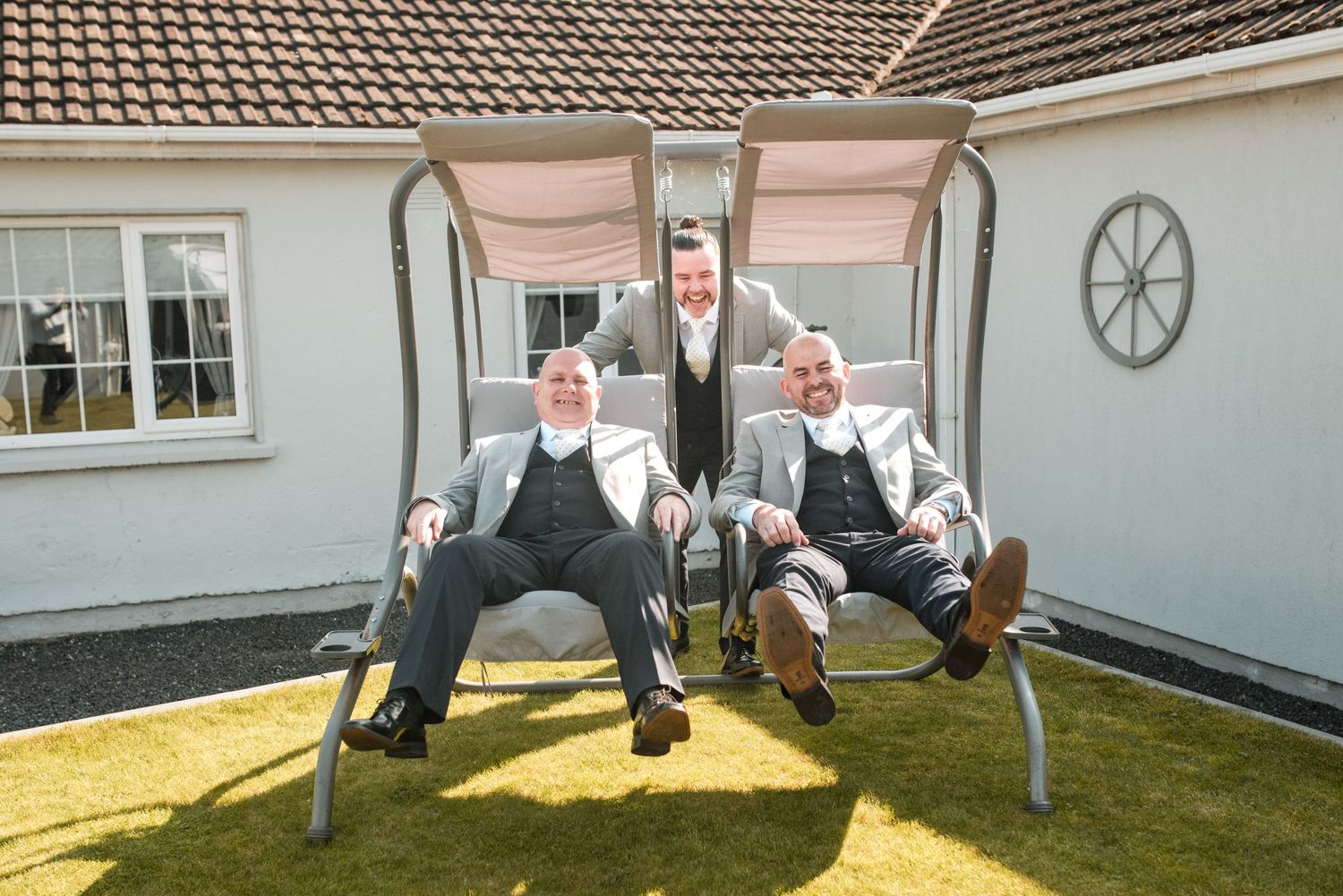 Three groomsmen in formal attire relaxing and posing playfully on an outdoor swing set in a backyard.