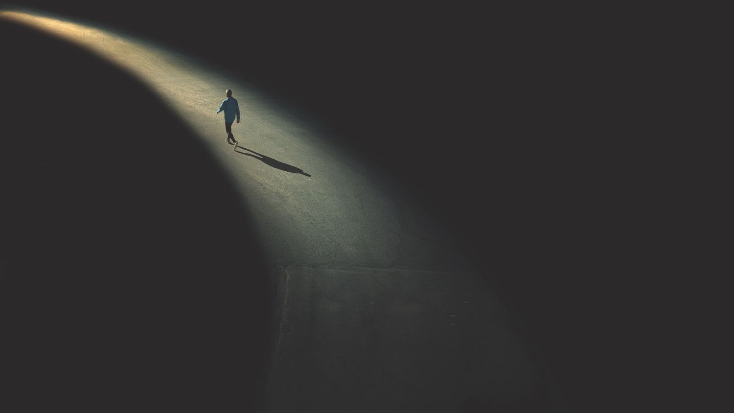 A silhouetted figure walks alone in a dark space illuminated by a single overhead light casting a long shadow.