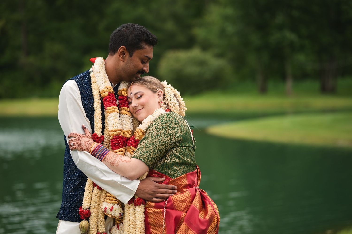 Couple in traditional German folk dress embrace romantically by a serene lake in a lush green setting.
