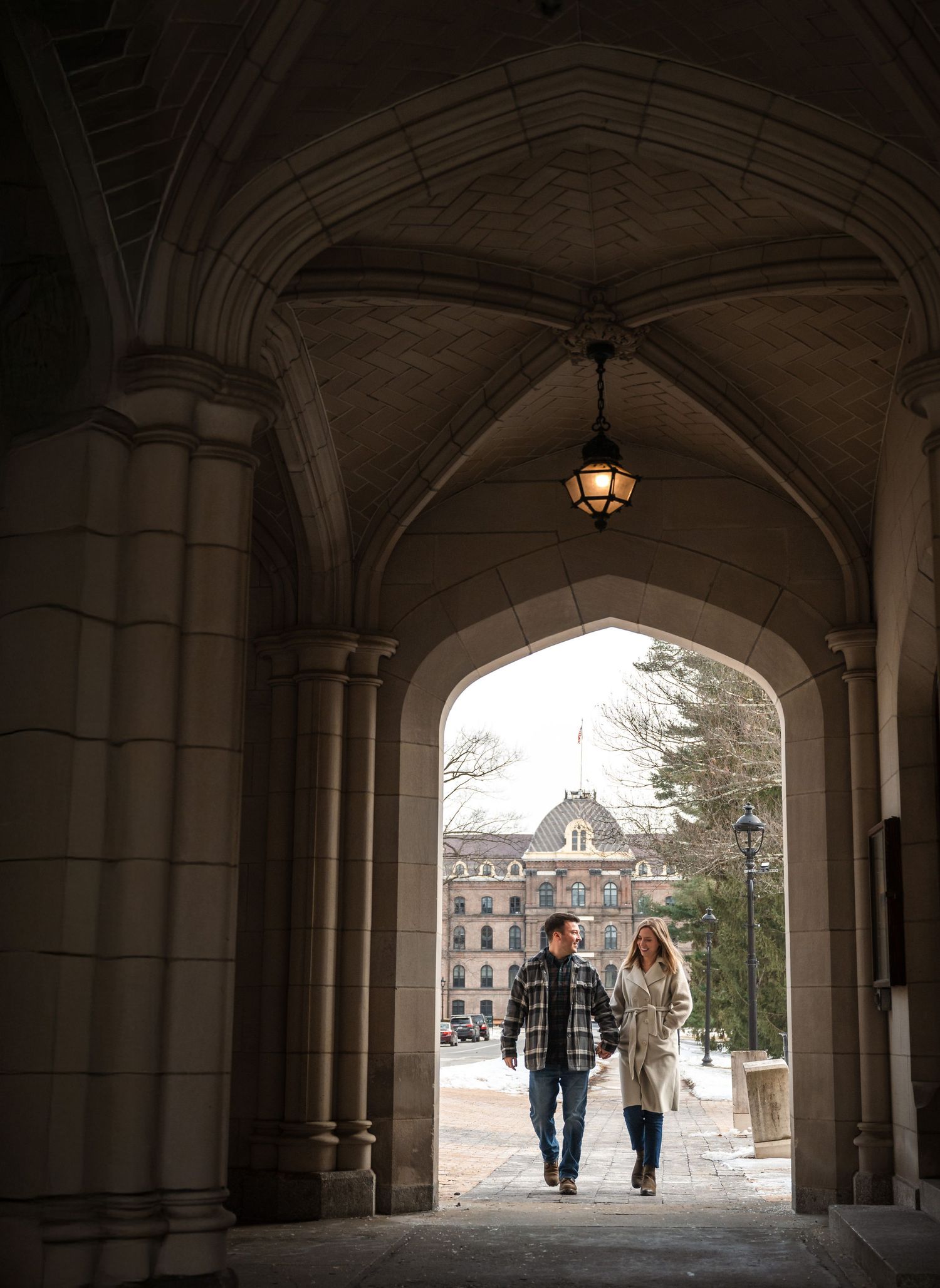Two students walk through a Gothic archway on a university campus with vaulted ceiling and pendant lighting.
