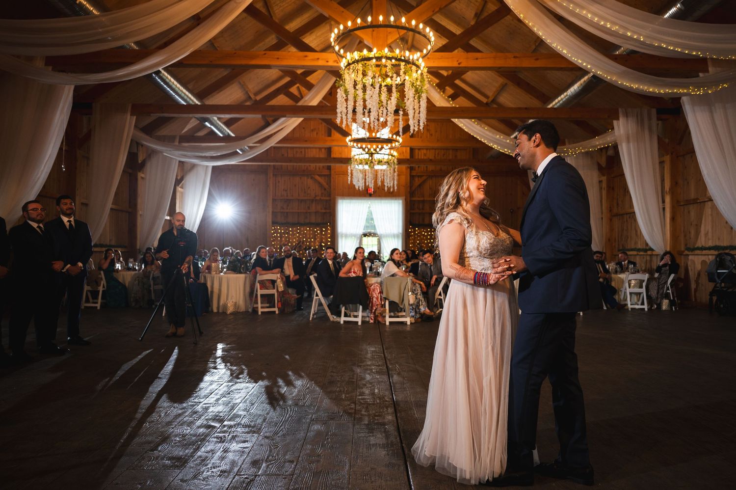 A couple dances at a rustic wedding reception inside an elegant barn with draped fabric and crystal chandeliers.