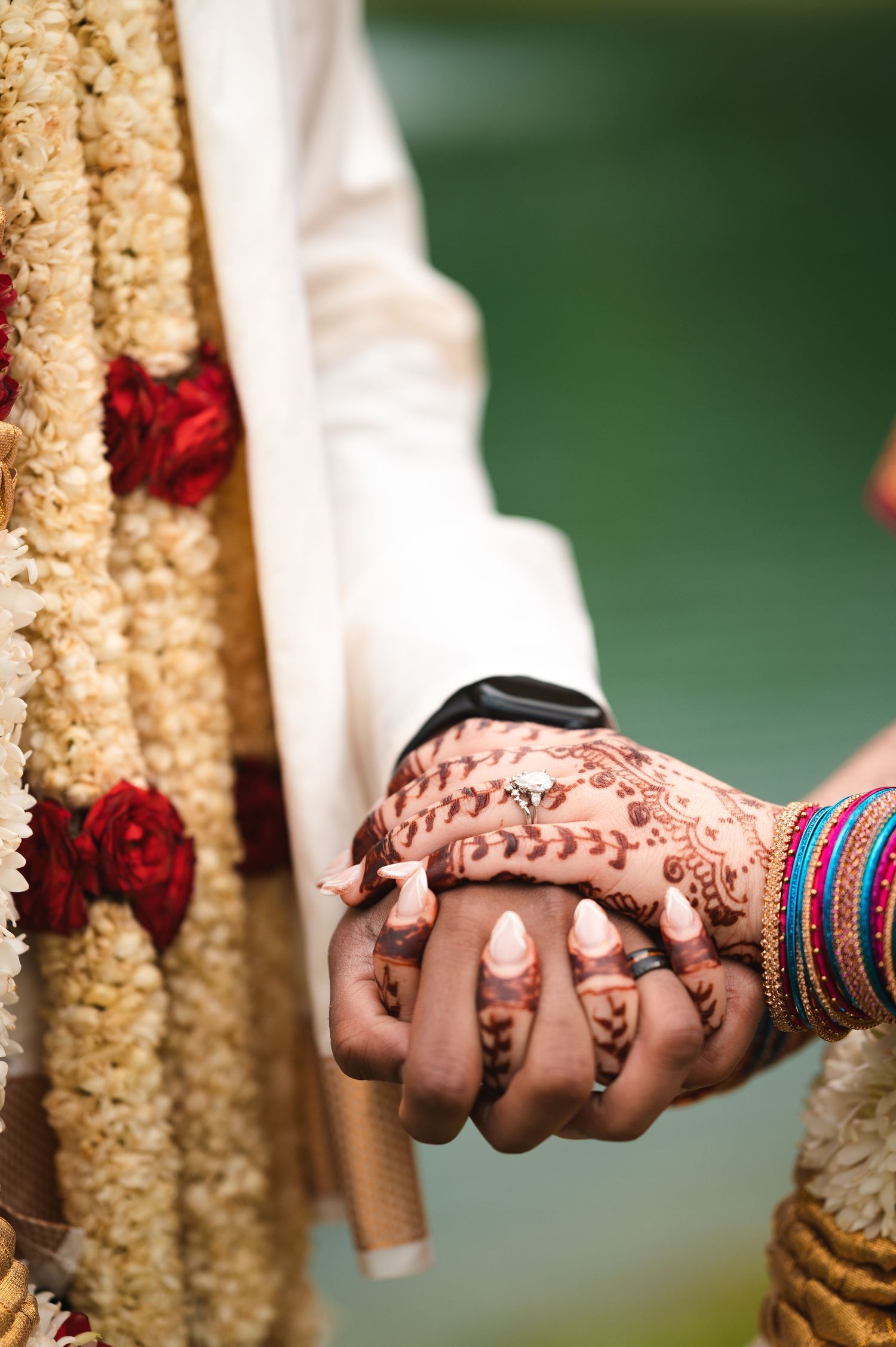 Clasped hands adorned with traditional henna and bangles during an Indian wedding ceremony against blurred floral backdrop.
