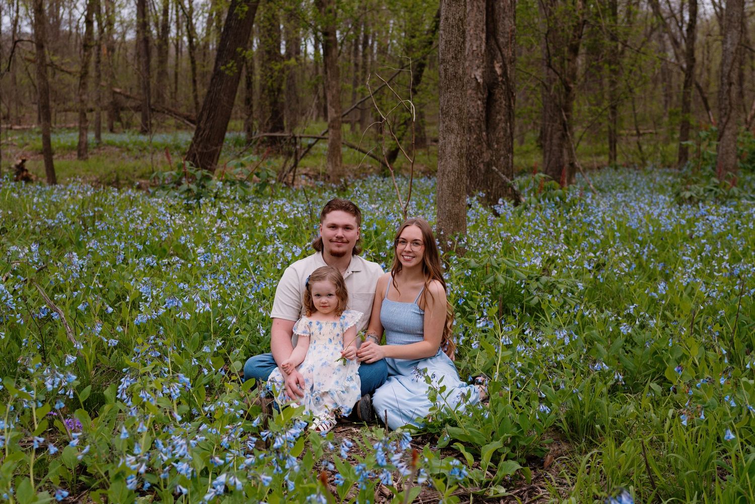 A family sits together among a field of blooming bluebells in a spring woodland setting.