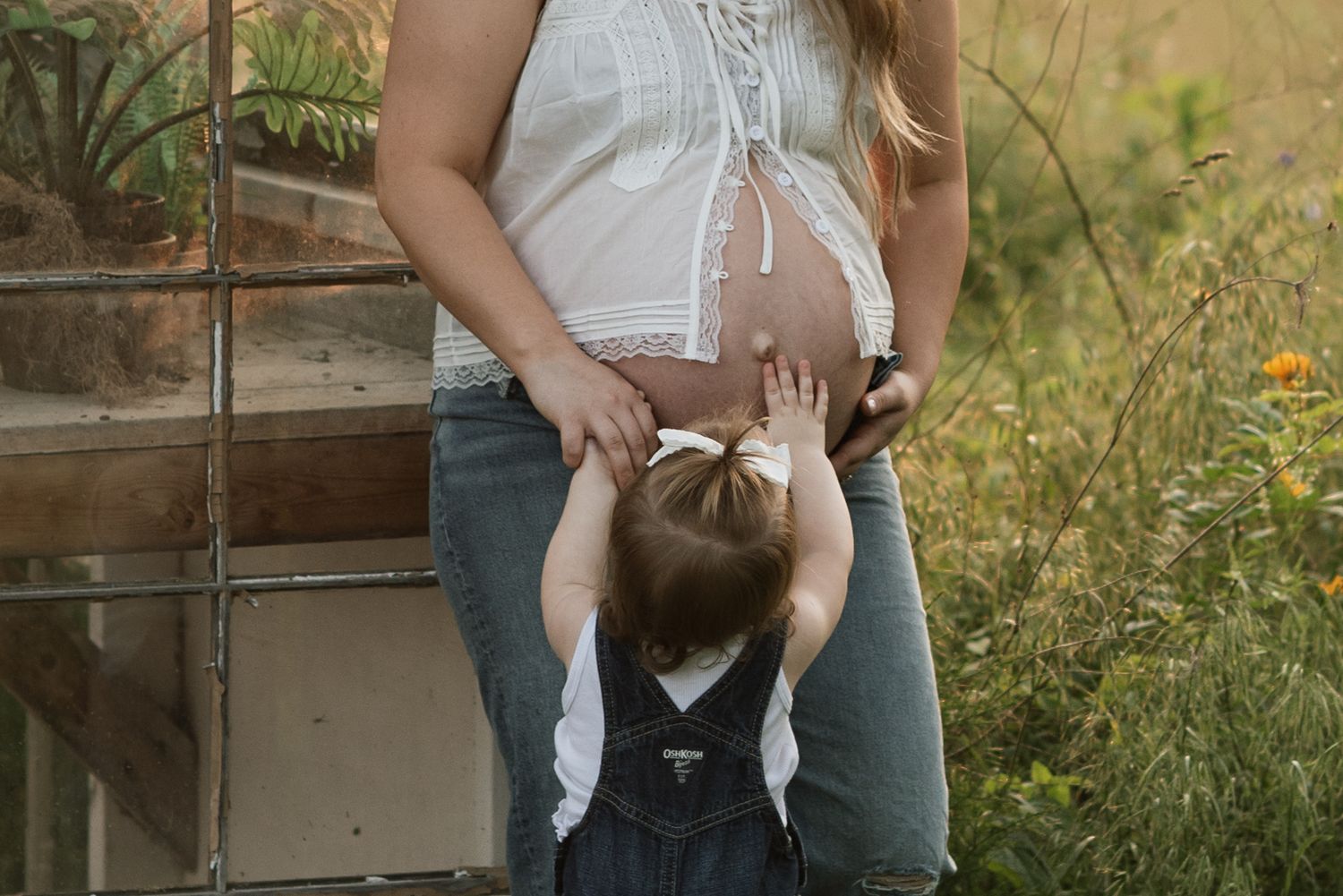 A toddler touches her mother's pregnant belly, surrounded by flowers in golden hour.