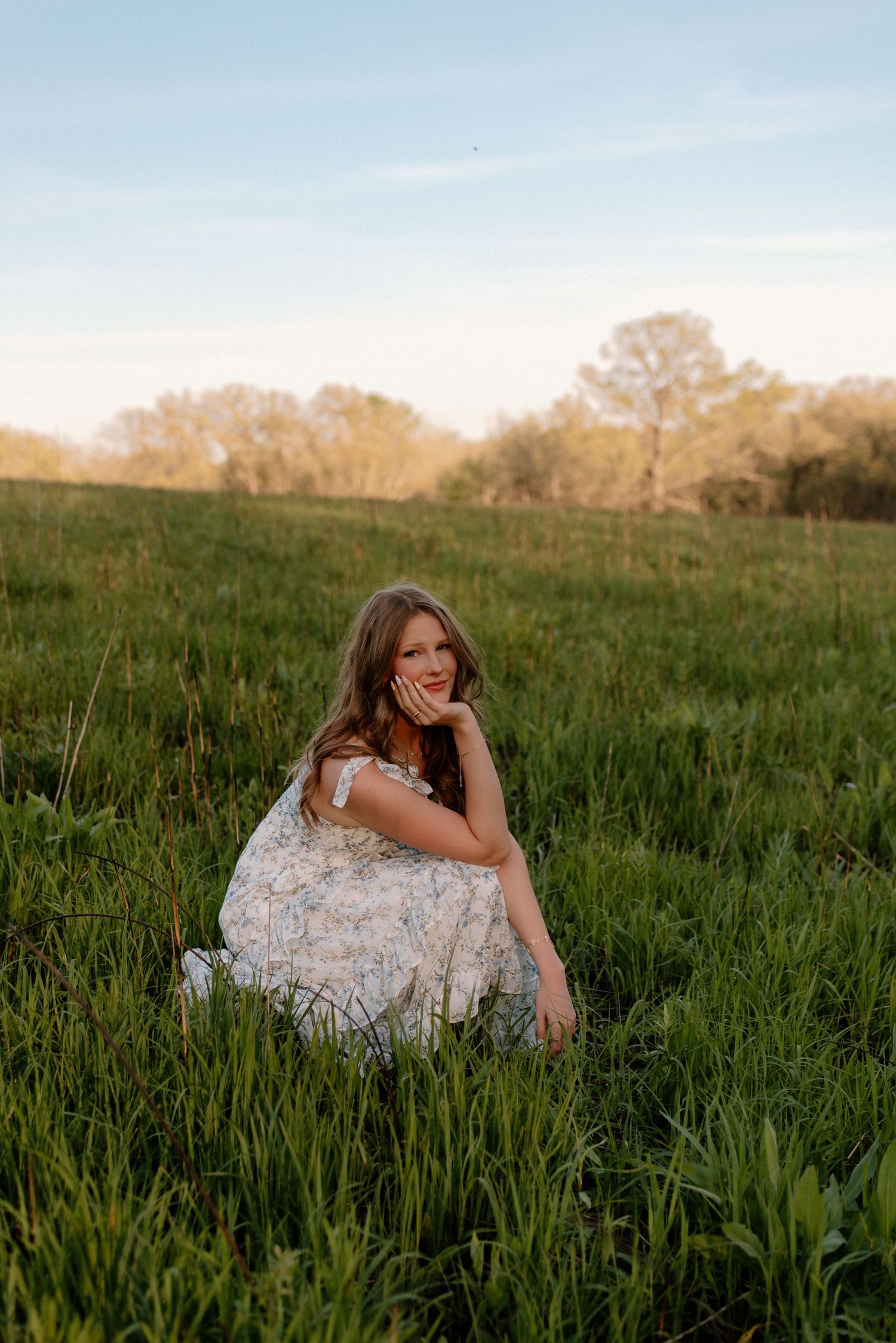 A girl in a flowing white dress reclines thoughtfully in a sunlit meadow of tall grass during golden hour.