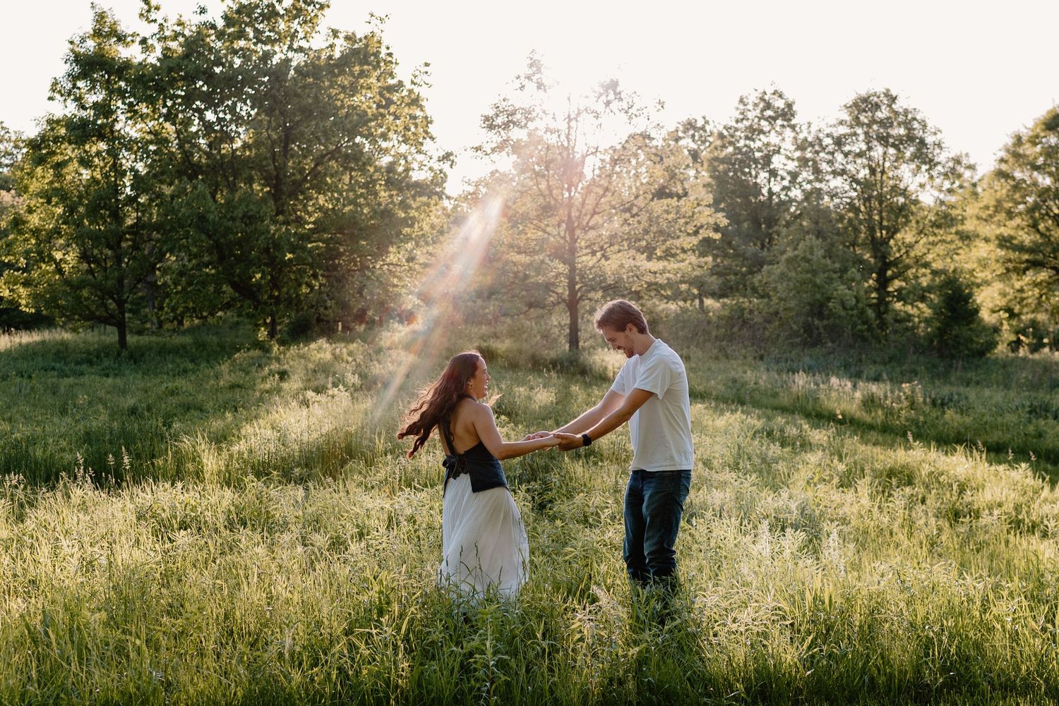A couple dances together in a sunlit meadow during golden hour while surrounded by trees and tall grass.