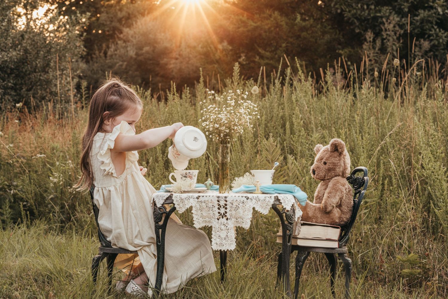 Young girl having outdoor tea party with teddy bear in sunlit meadow.