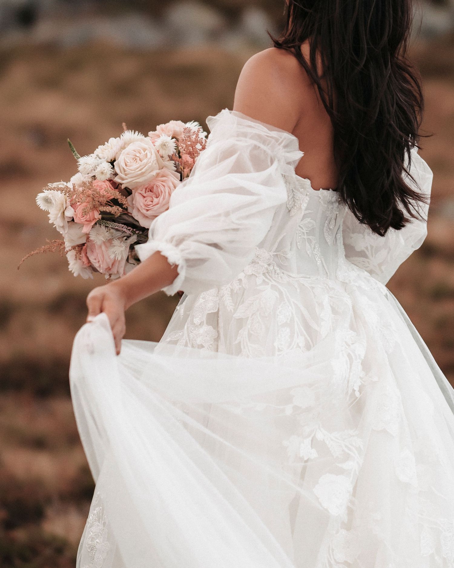 A flowing white wedding dress and blush pink bouquet are showcased against a rustic outdoor backdrop.