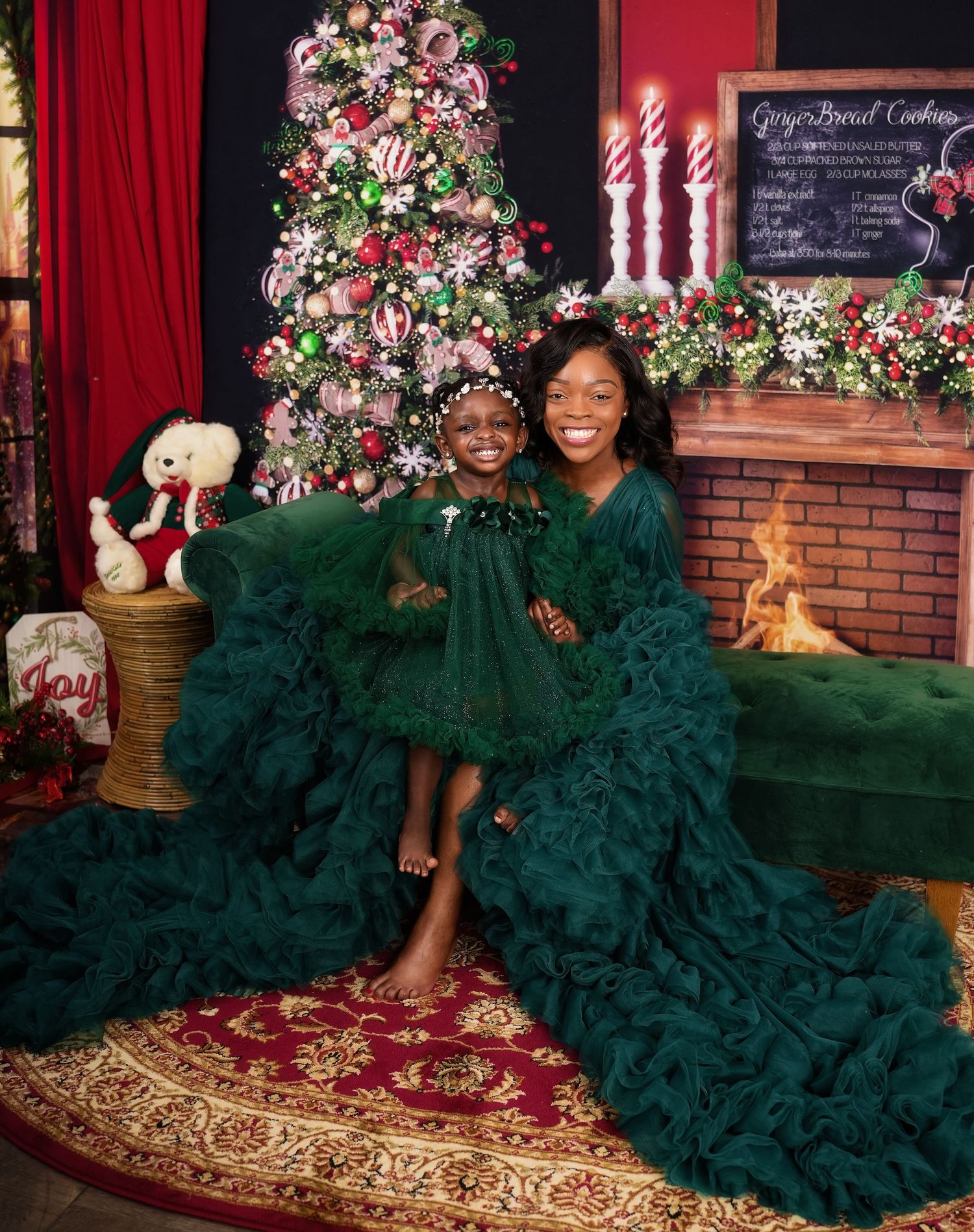 Two people wearing matching emerald green tulle gowns pose together on red carpet for holiday photos against brick backdrop.