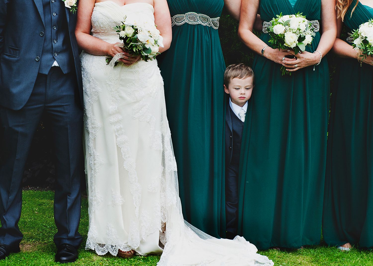 A young boy in a suit peeks out playfully from between the bridesmaids' dresses, adding a humorous touch to the otherwise formal scene.