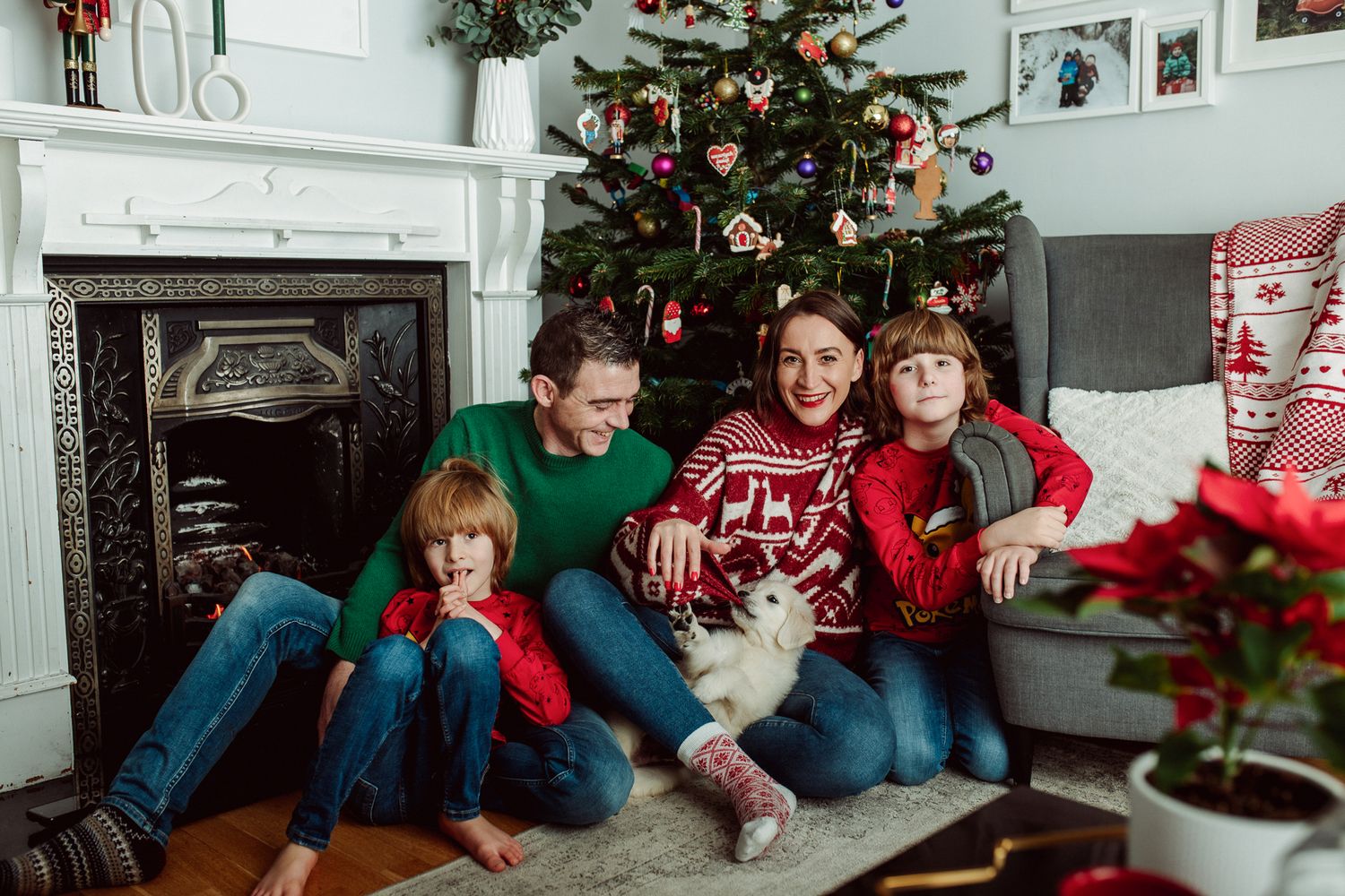 A family in festive clothing sits comfortably by a decorated Christmas tree, with a playful dog nestled among them.