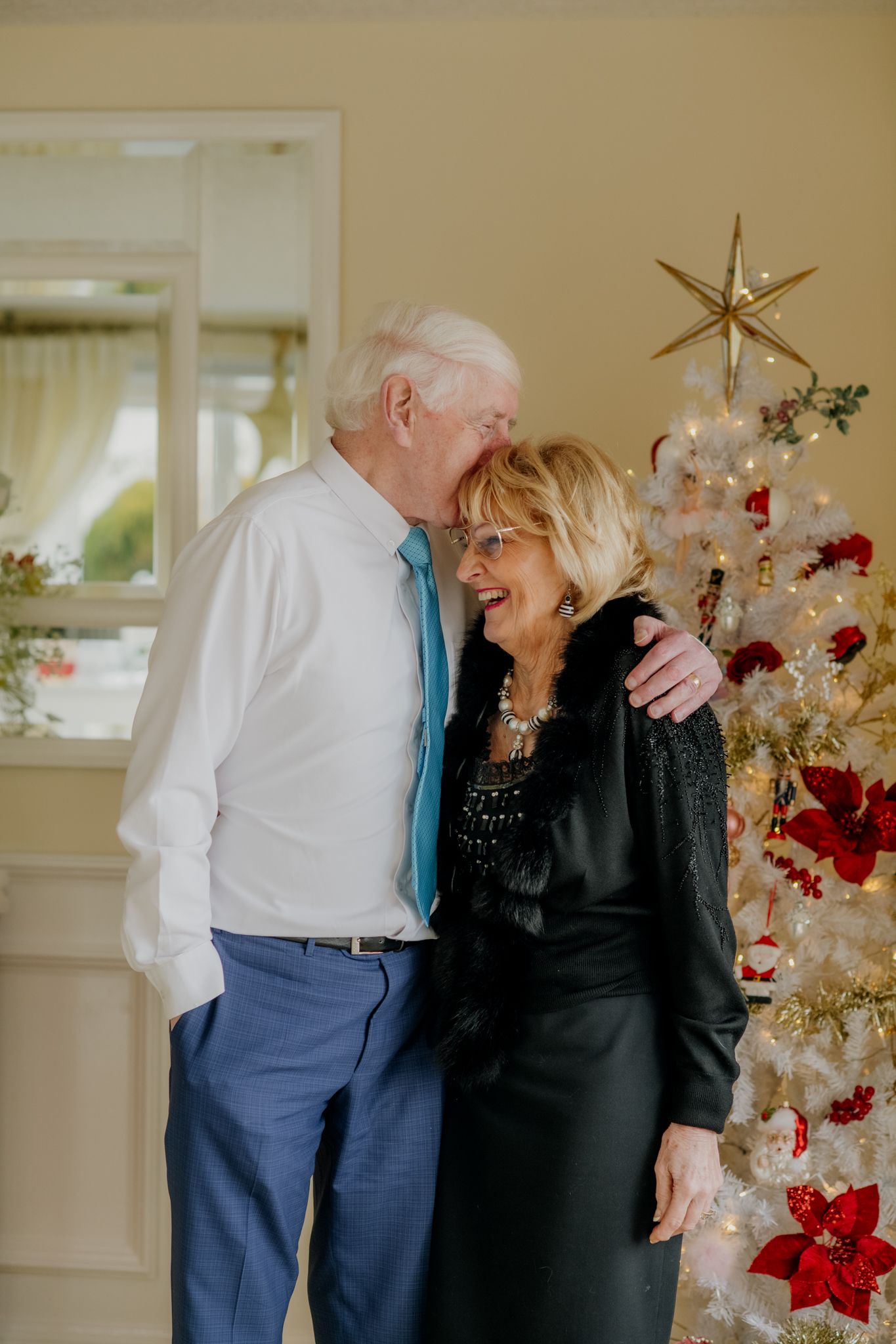 A couple embraces affectionately in front of a decorated Christmas tree, dressed elegantly for a festive occasion.
