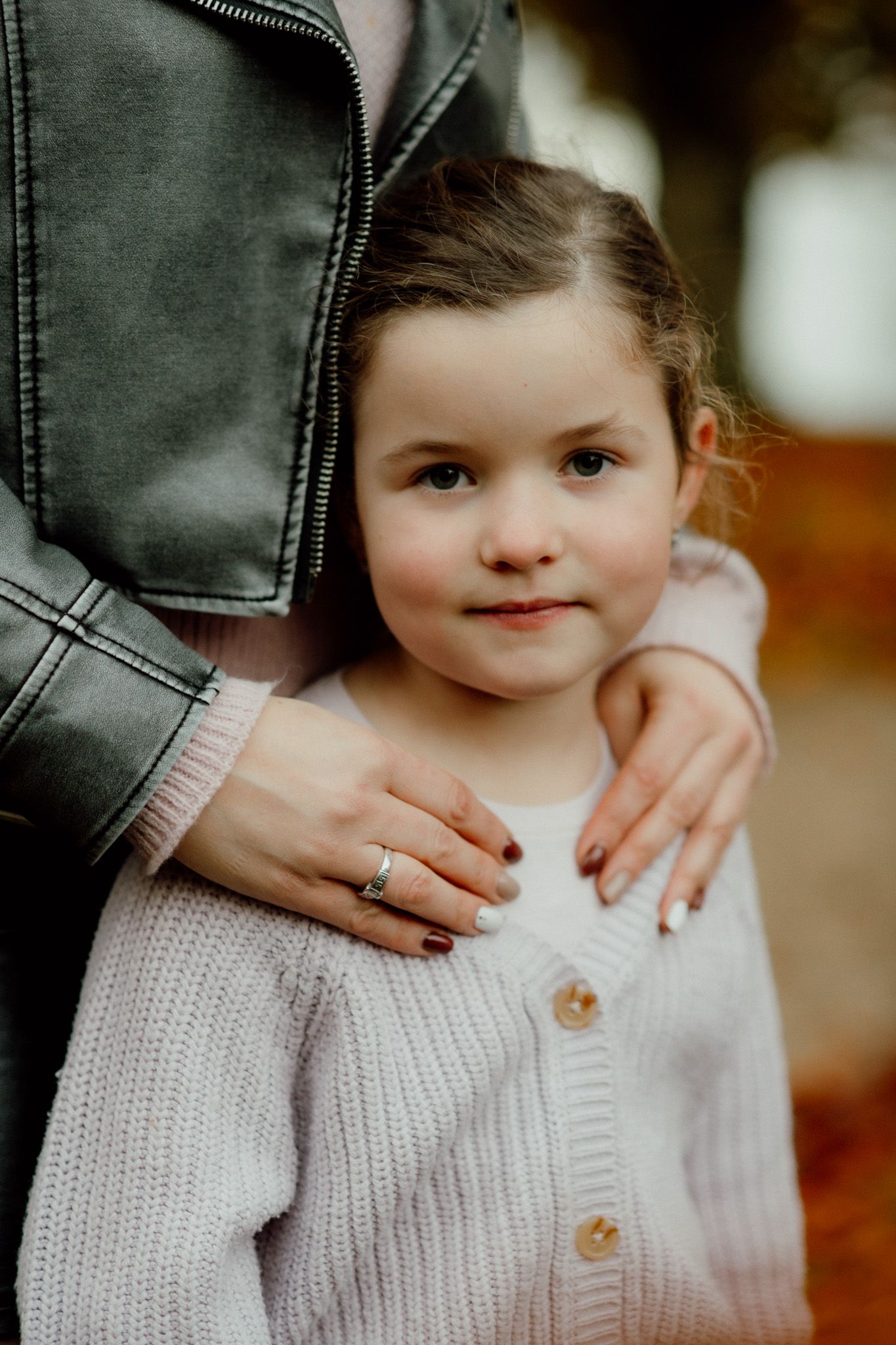 A close-up of a woman’s hands gently resting on a child wearing a light purple knitted cardigan, set against a blurred autumn backdrop.