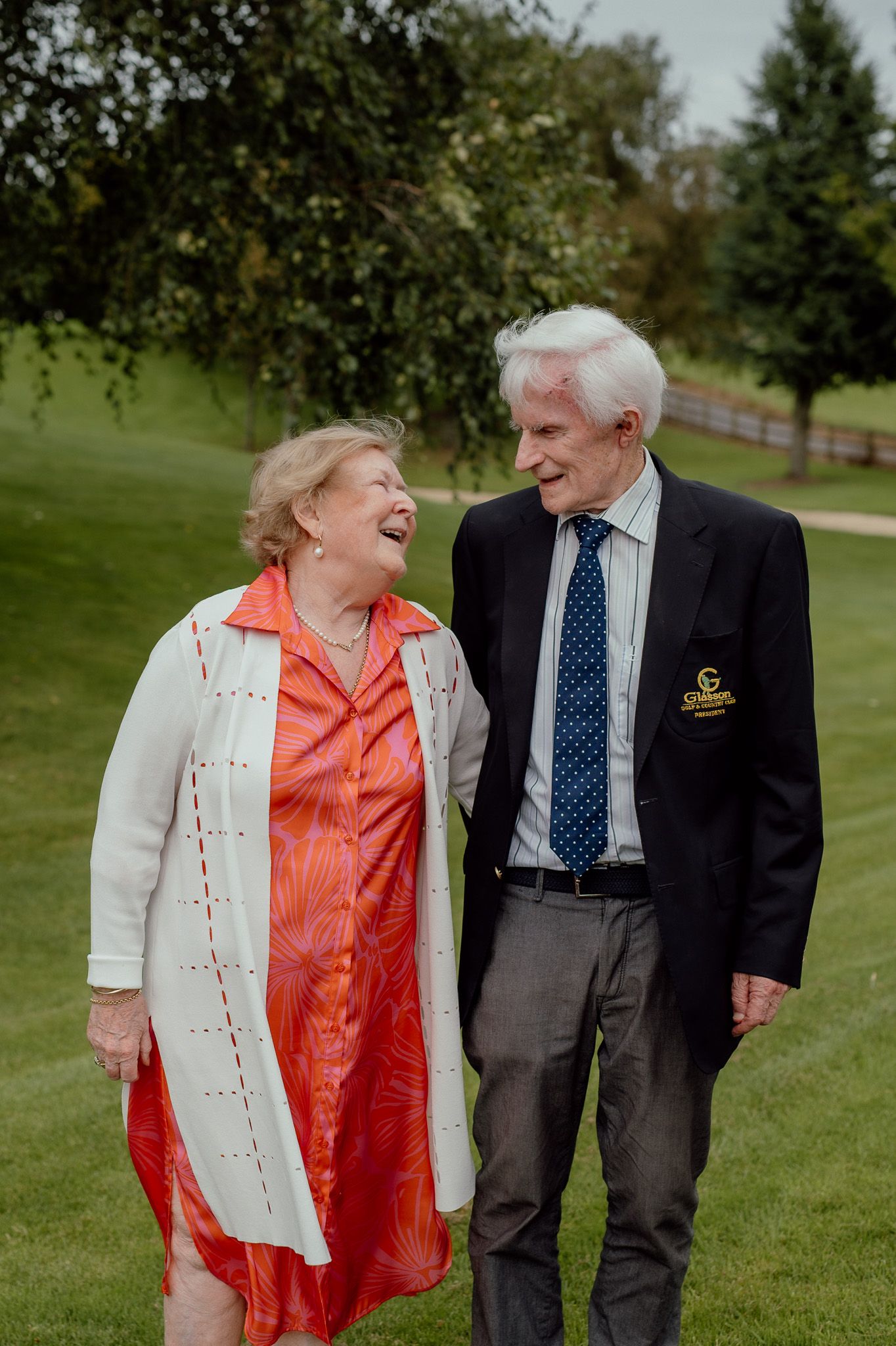 An elderly couple stands hand in hand on a green lawn, wearing colorful outfits and smiling gently at the camera.