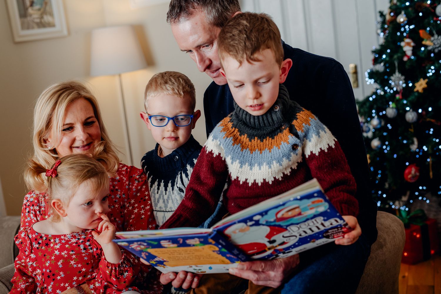 A cozy indoor scene with children gathered around an adult reading a festive book, with a decorated Christmas tree in the background.