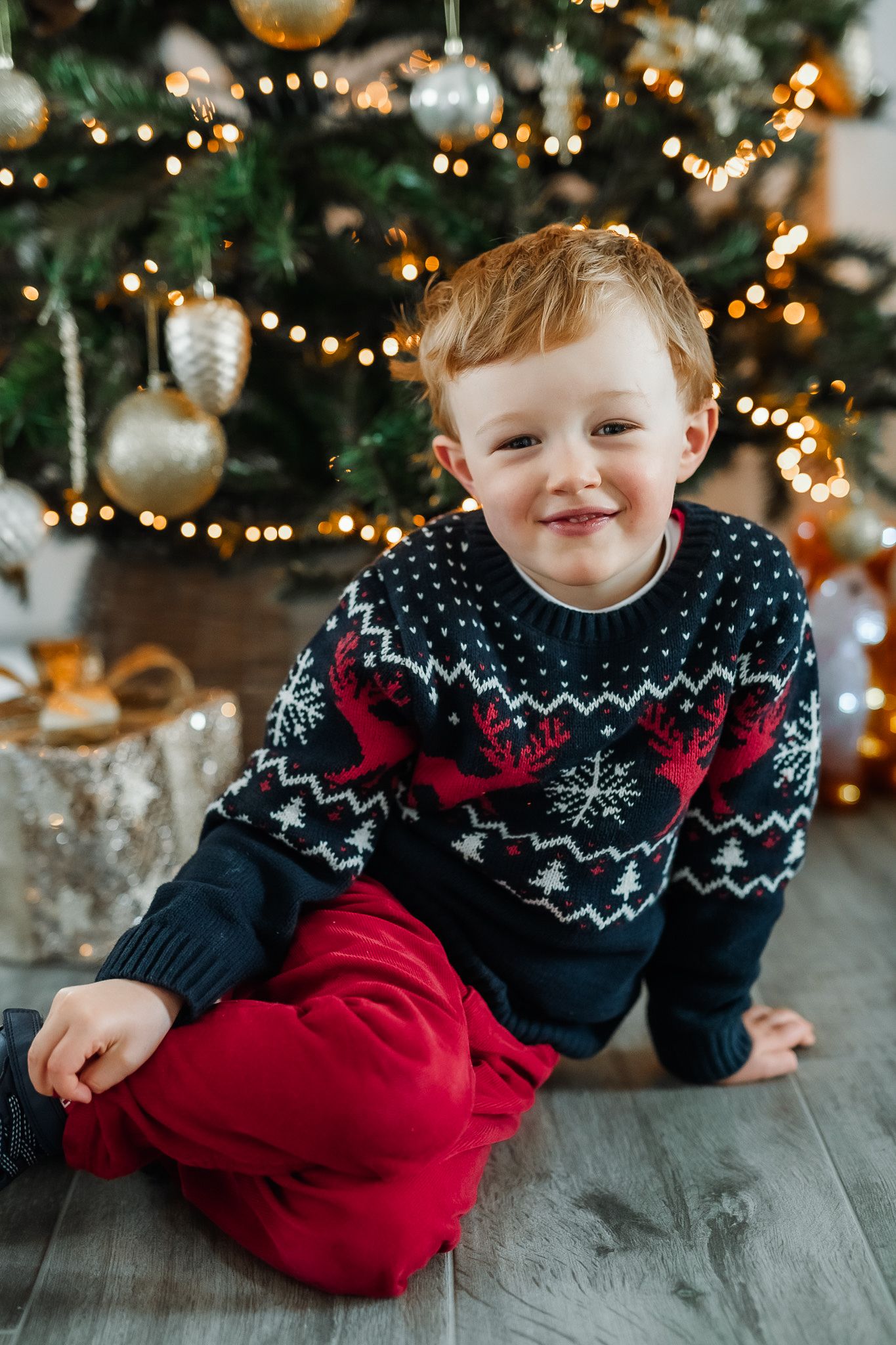 A child in a festive sweater sits on the floor near a Christmas tree adorned with golden ornaments and twinkling lights.