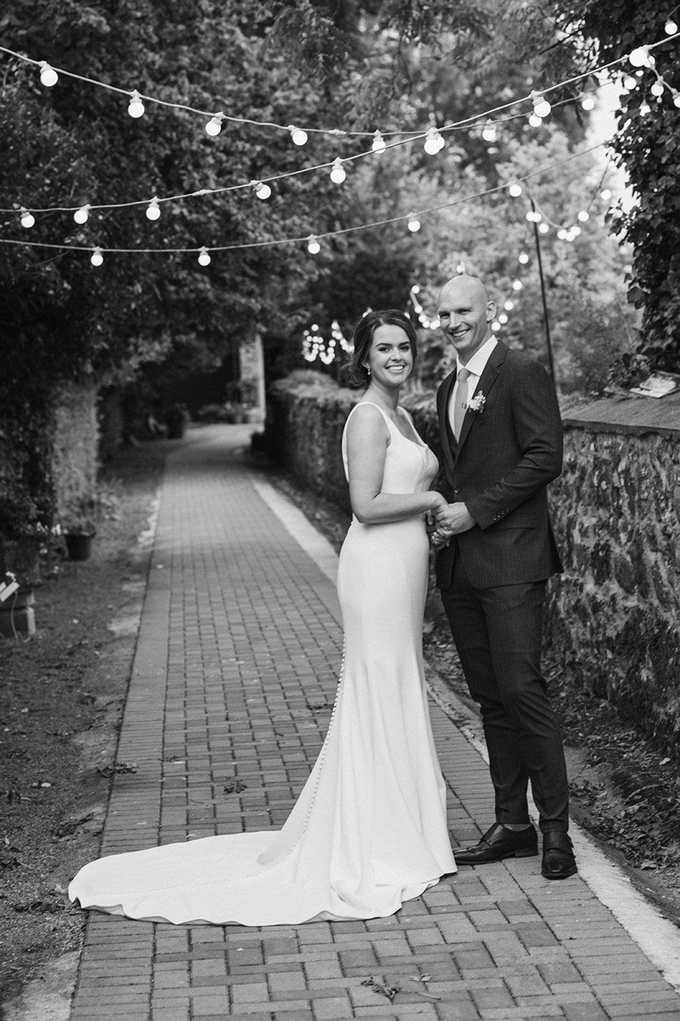 A black and white photograph of a newlywed couple captures them standing hand in hand on a charming brick pathway at The Barn, Tyrrellspass.