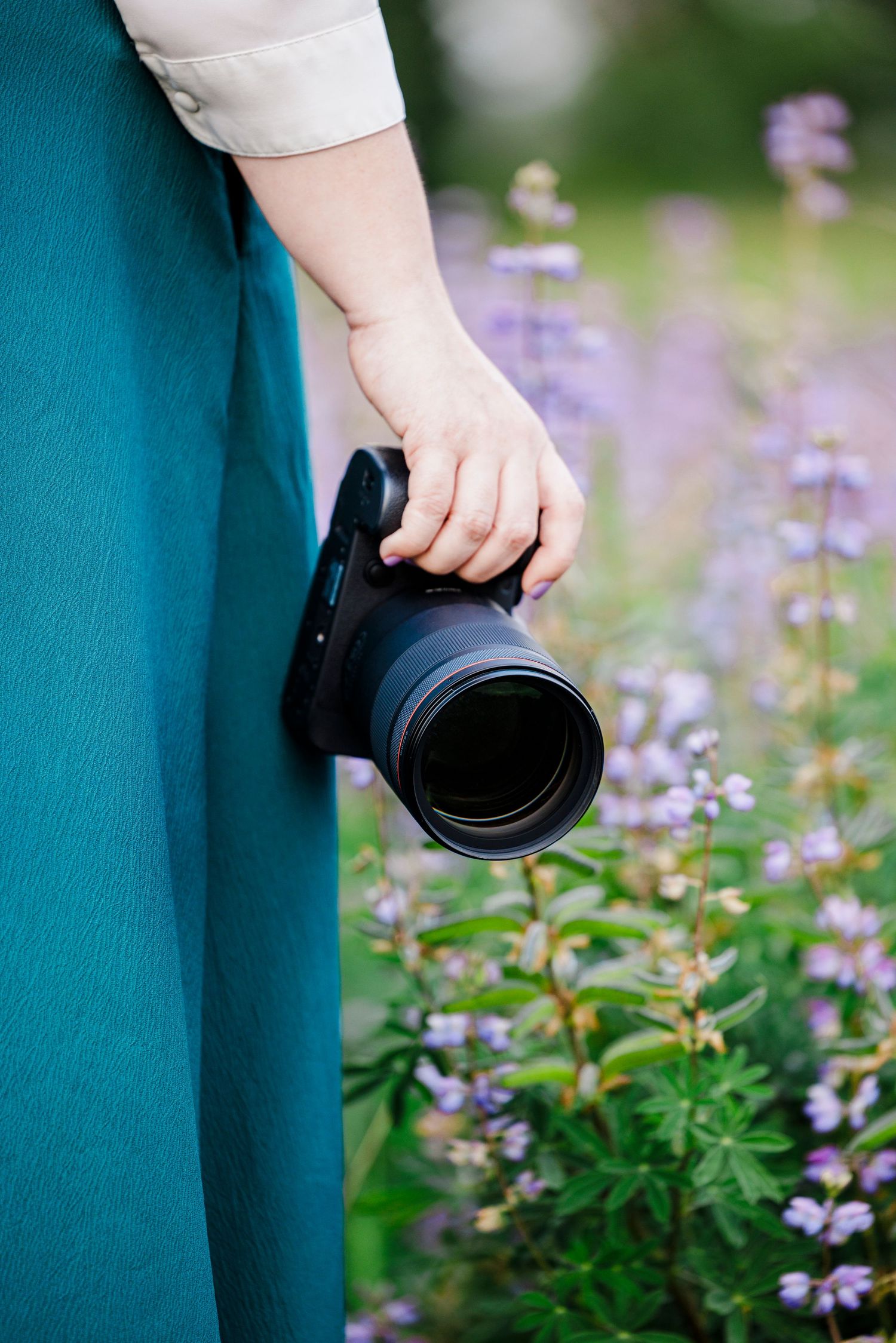 Hand holding a professional camera lens against backdrop of blooming purple wildflowers and teal dress.