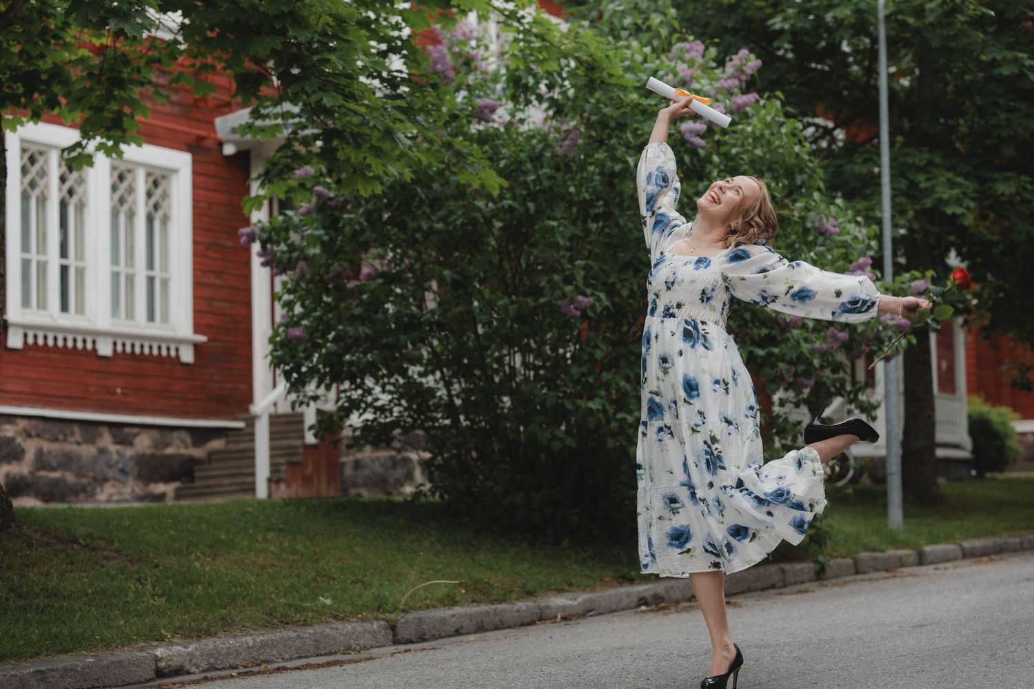 A woman in a white floral dress twirls playfully on a quiet residential street with brick buildings in the background.