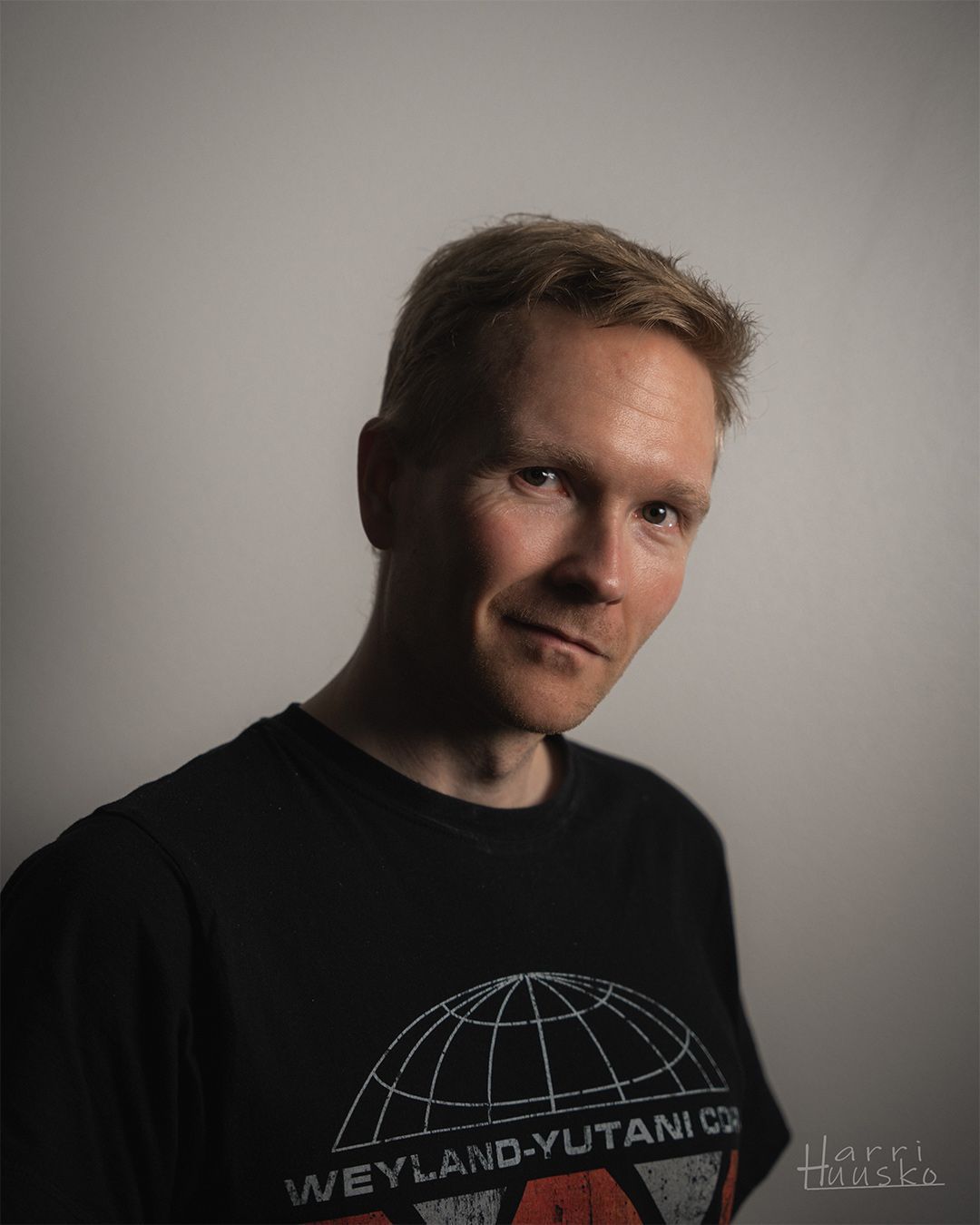 Portrait shot against grey background of someone wearing a black t-shirt with Weyland-Utani Co logo design.