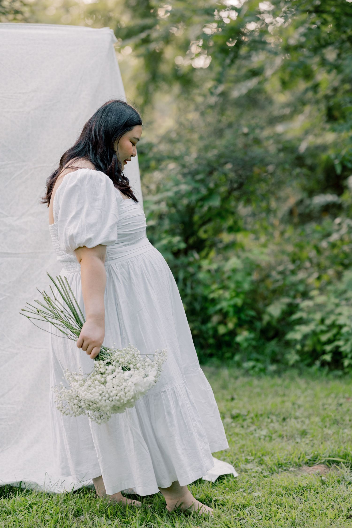 A person in a flowing white dress holds a delicate bouquet of baby's breath flowers in a garden setting.