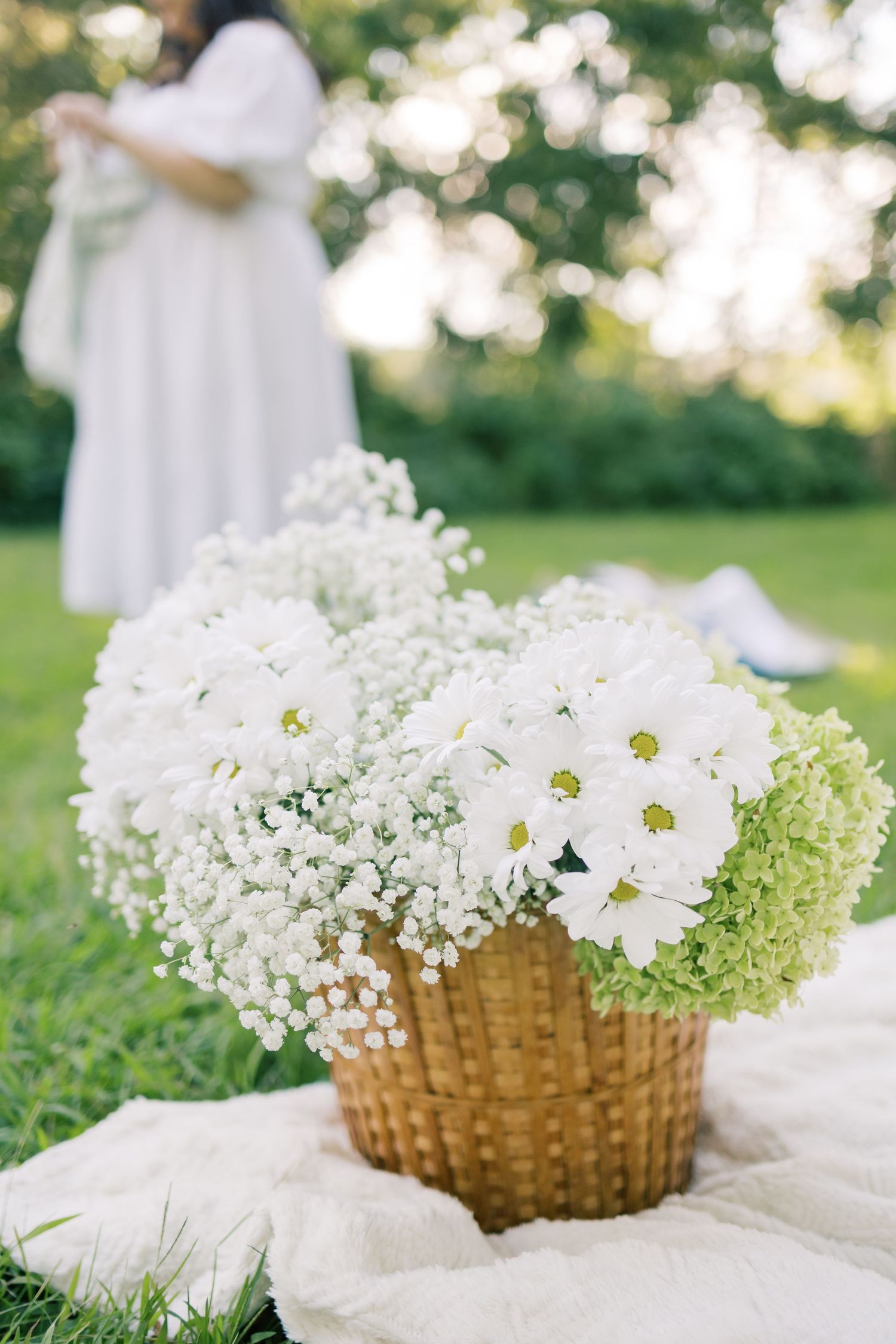A woven basket filled with white daisies and baby's breath flowers sits on a blanket in a sunny garden.