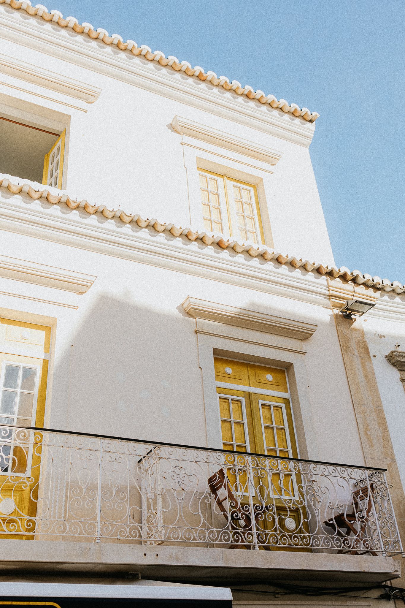 Traditional white Mediterranean building with yellow doors and windows featuring an ornate wrought iron balcony against blue sky.