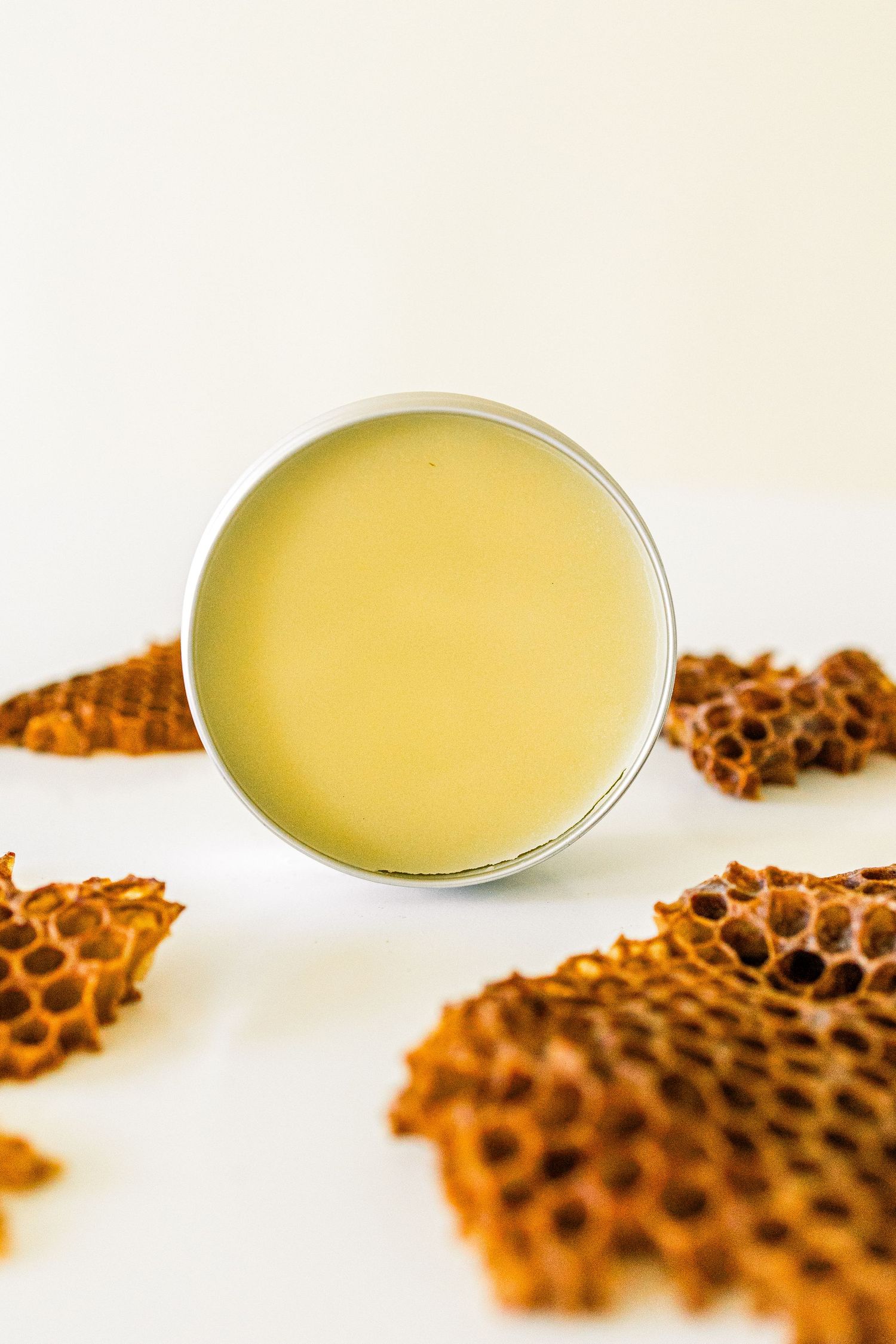 A tin of yellow beeswax balm or salve surrounded by decorative honeycomb pieces on a white background.