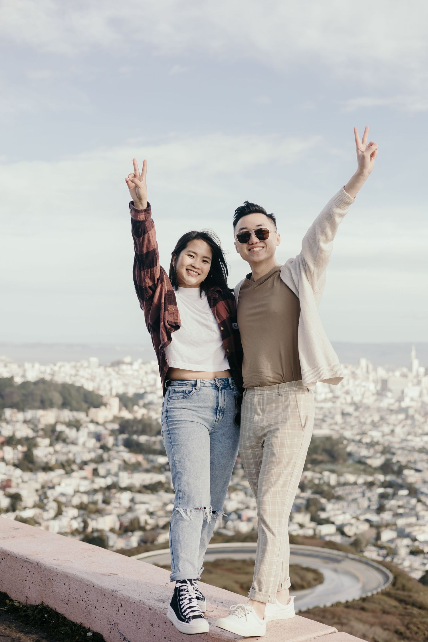 Two tourists making peace signs while posing together on scenic overlook with cityscape view in background.