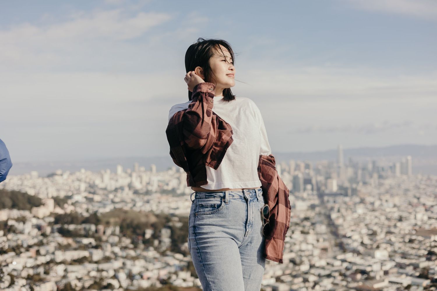 Someone stands on a hilltop overlook wearing jeans and a white sweater with a cityscape view in the background.