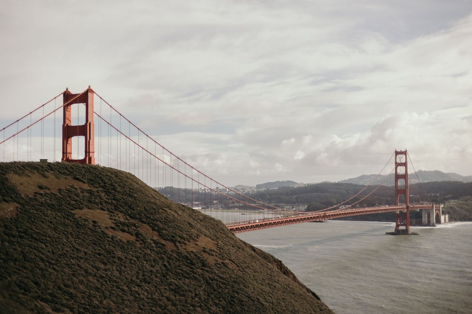 Iconic Golden Gate Bridge spans San Francisco Bay under cloudy skies with rolling hills in the foreground.
