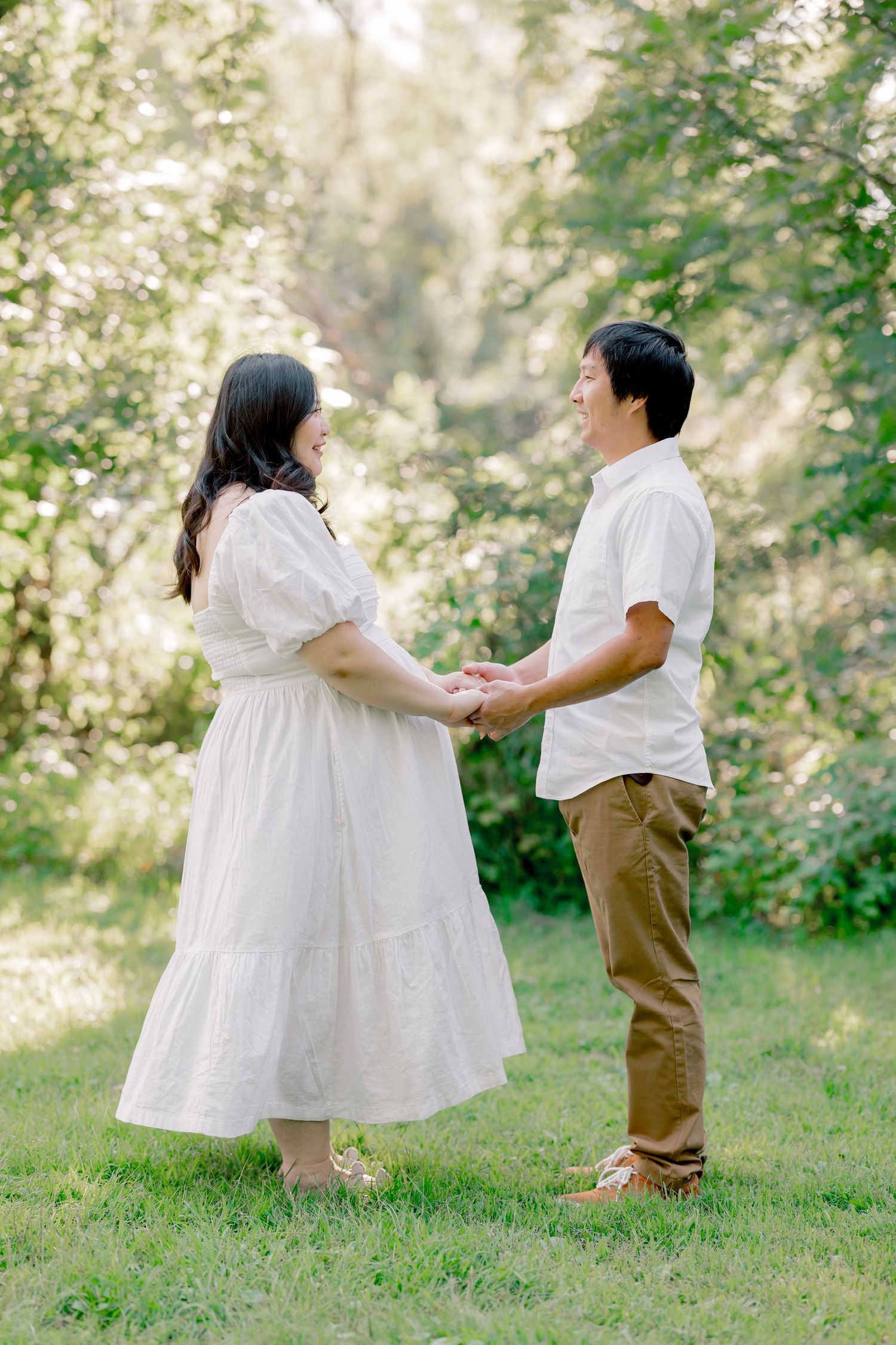 A couple holds hands in a sunlit garden, wearing coordinated white and khaki outfits against a natural backdrop.