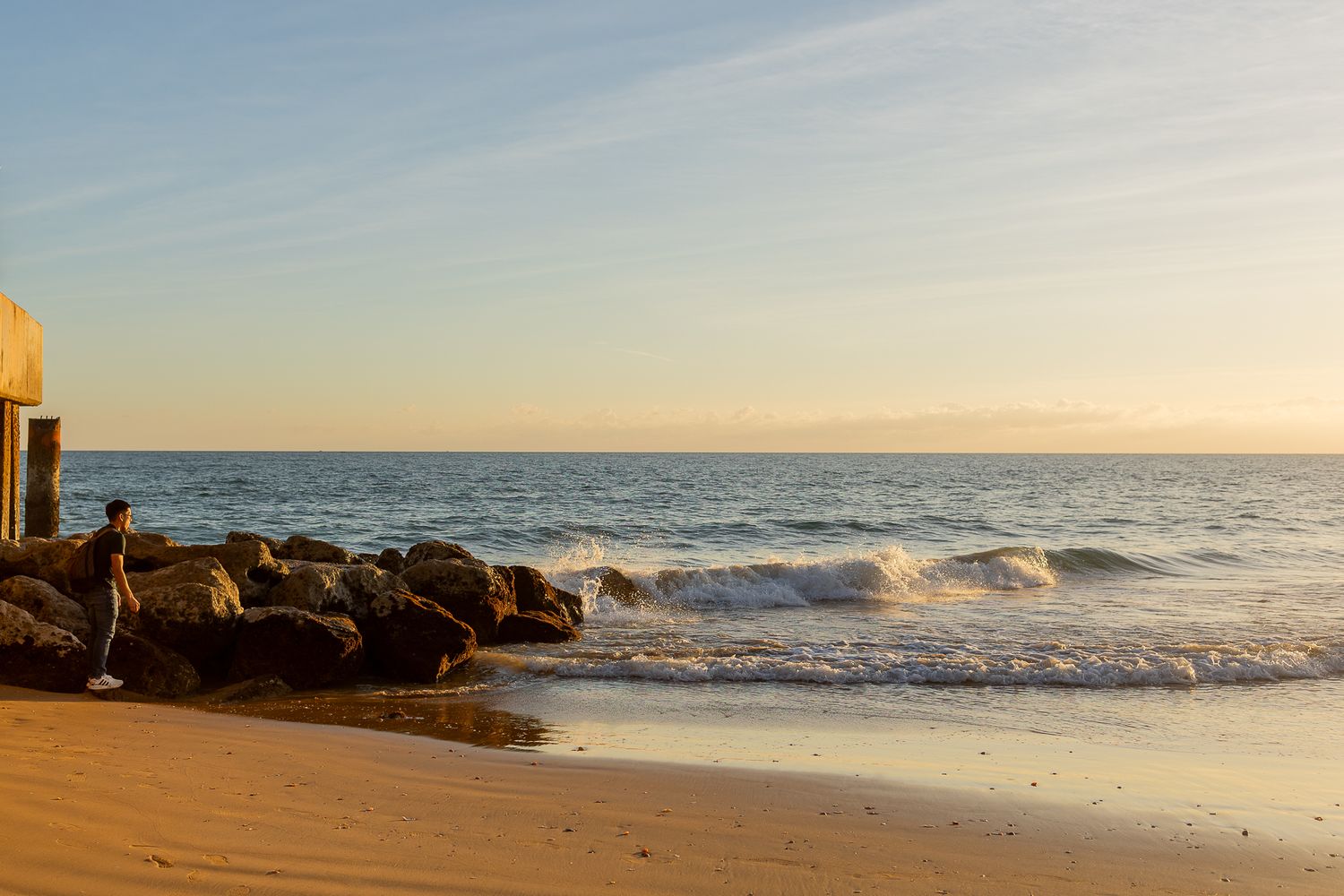 Rocky beach shoreline at sunset with gentle waves rolling onto golden sand under a warm sky.