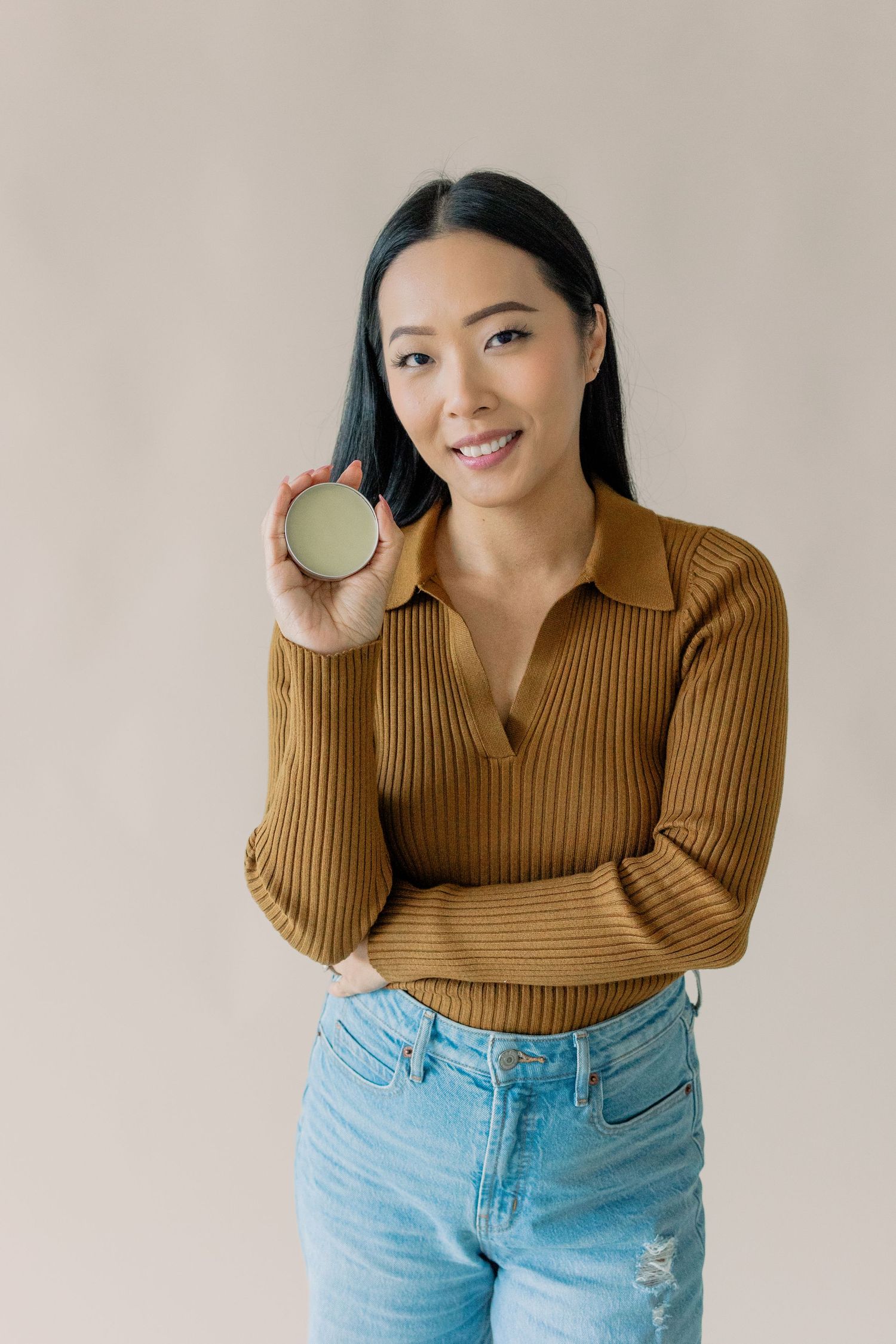 A woman in a brown ribbed sweater and light jeans holds up a round beauty product while standing against a beige wall.