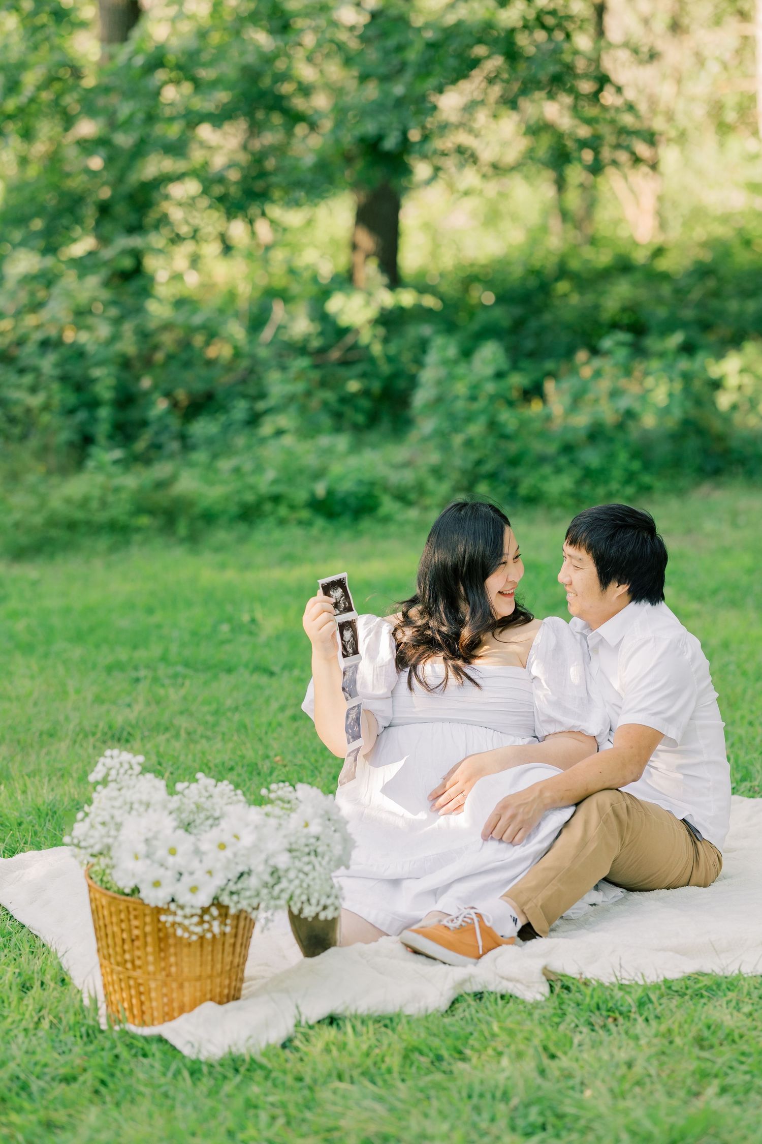 A couple enjoys a romantic picnic in a sunny park with a wicker basket of white flowers beside them on a blanket.