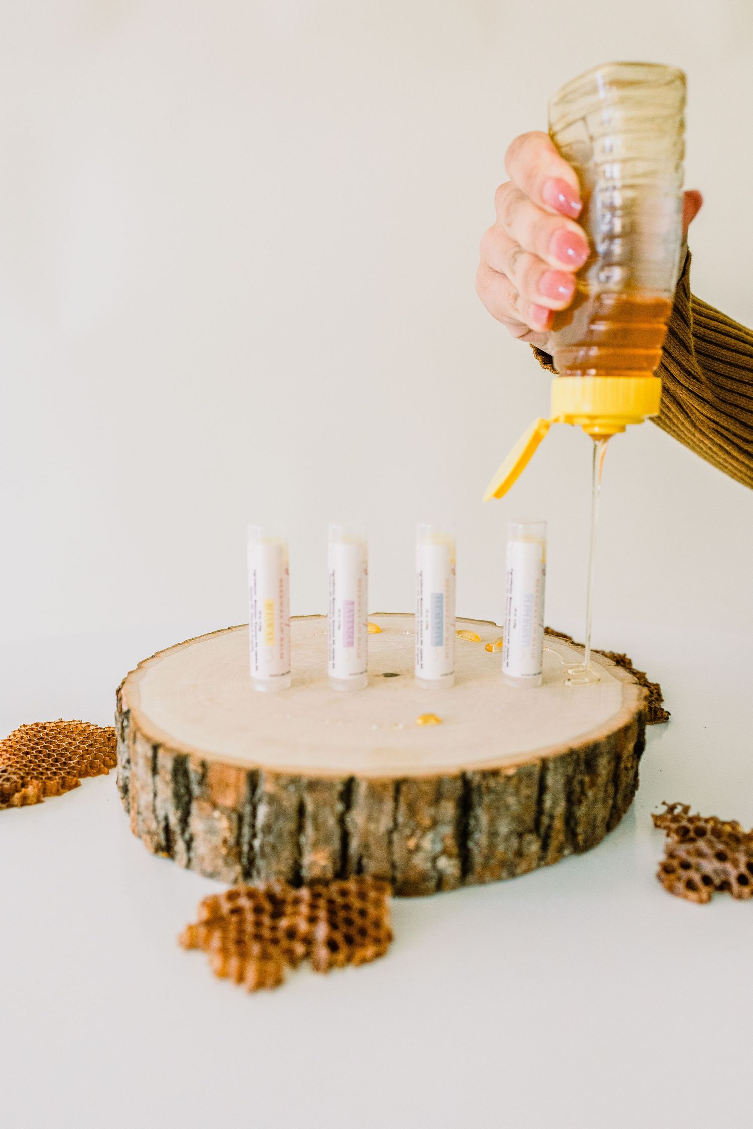 Hand pouring honey onto test tubes displayed on a rustic wood slice with honeycomb pieces as decoration.