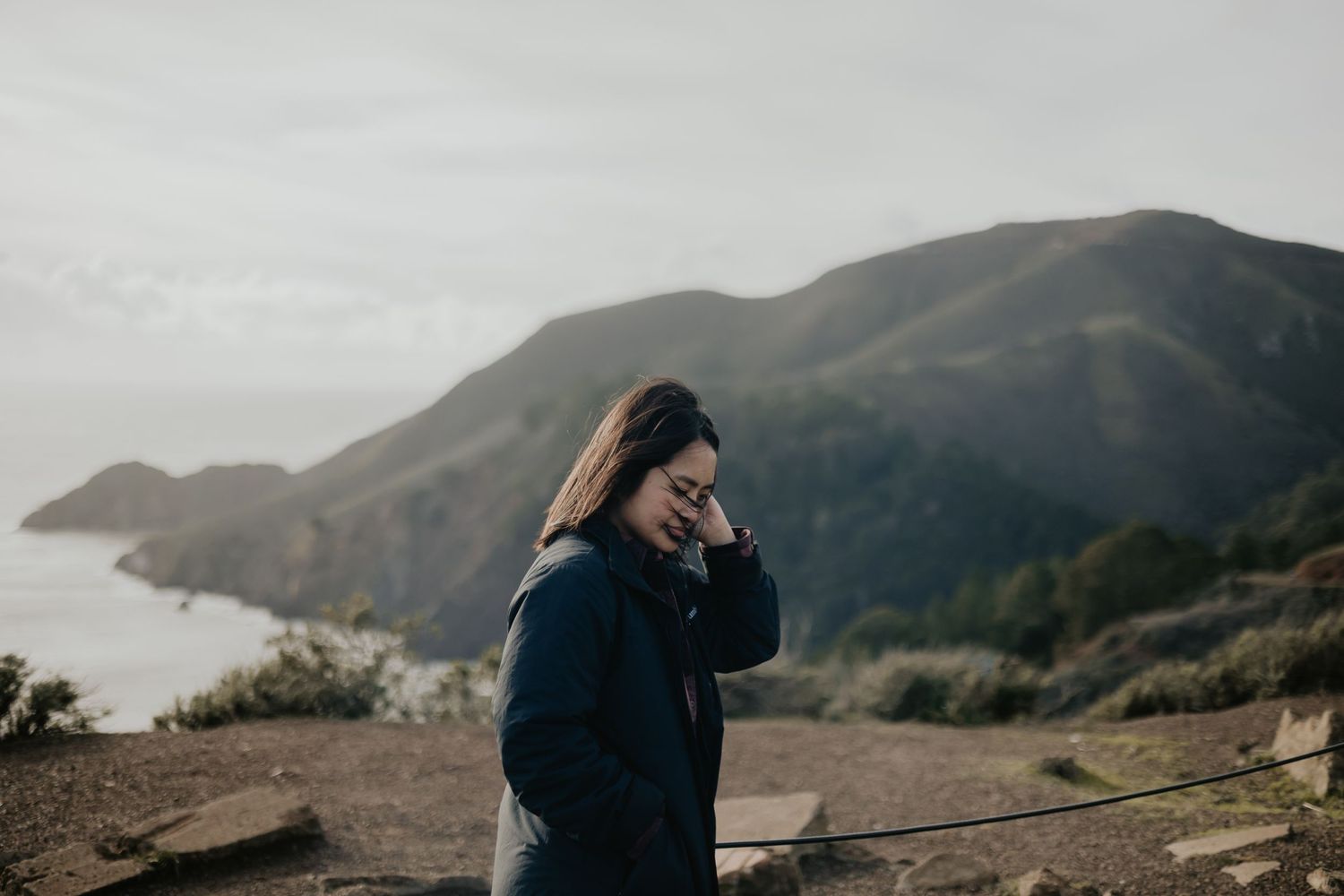 Someone in a blue jacket stands on a coastal cliff overlooking mountains and ocean on a cloudy day.