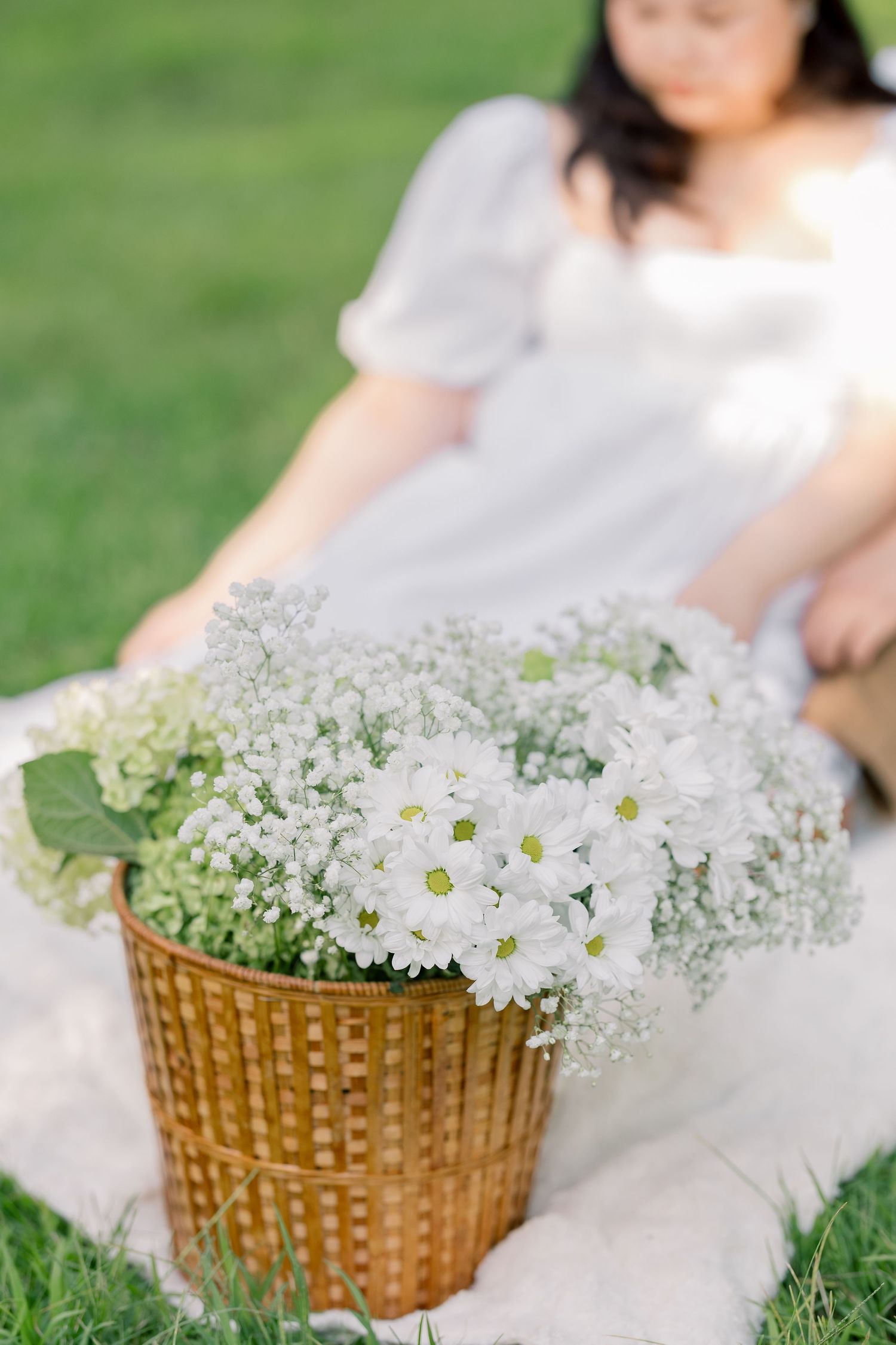 A wicker basket arrangement of white daisies and baby's breath flowers on grass.
