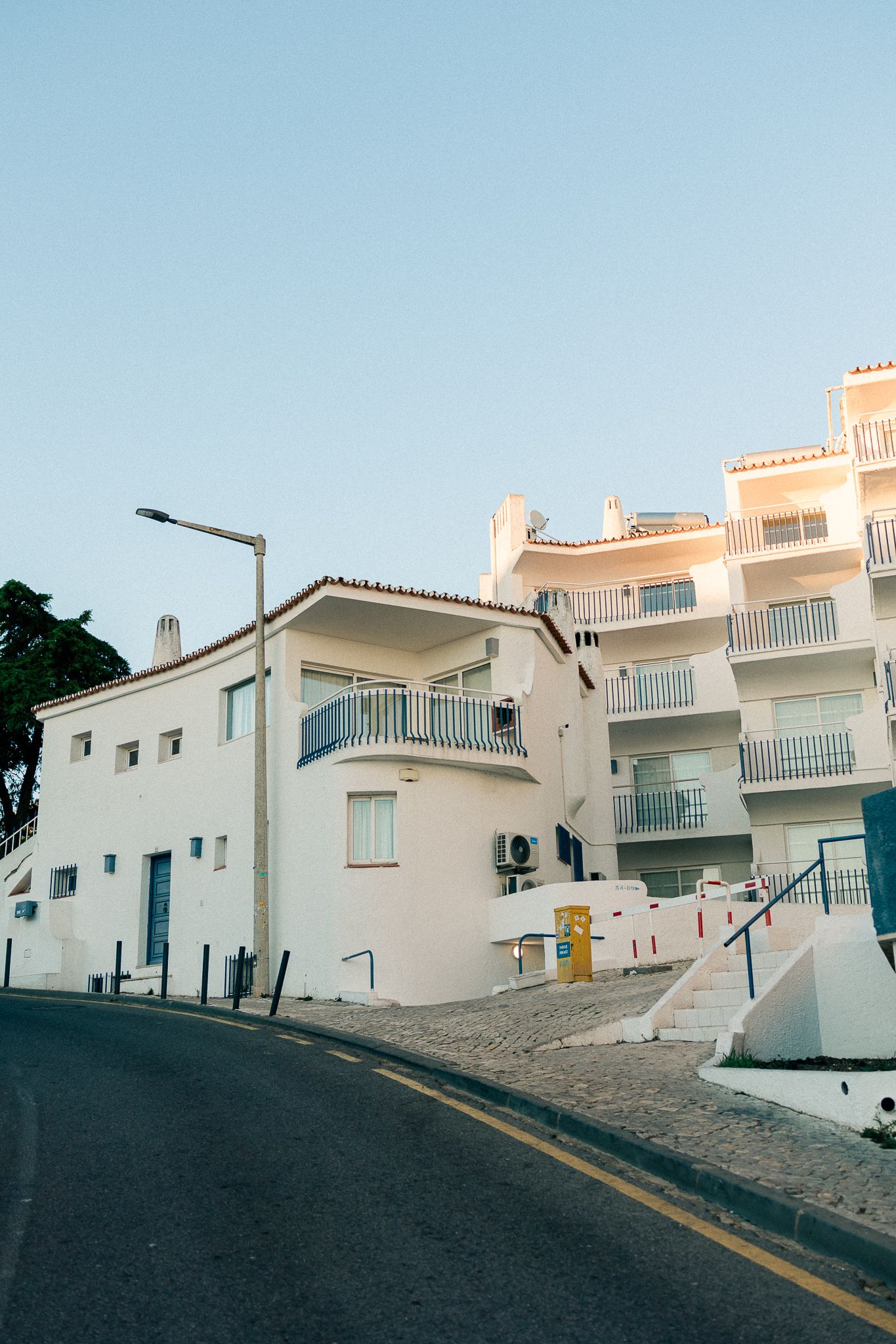 A curving hillside road leads to white and pink Mediterranean-style apartment buildings with balconies under a blue sky.