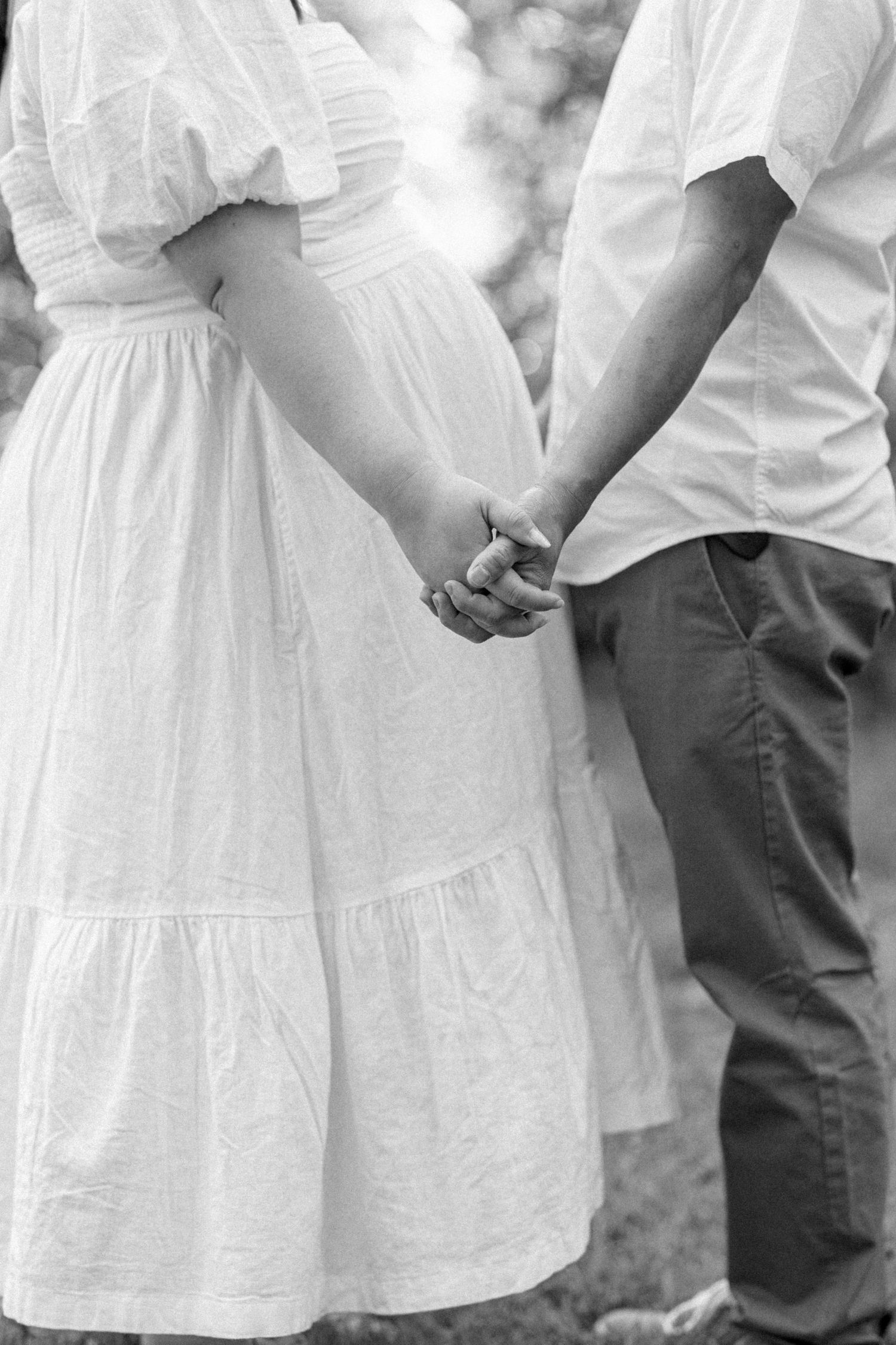 A couple holds hands tenderly while wearing white dress and casual attire in a black and white romantic photograph.