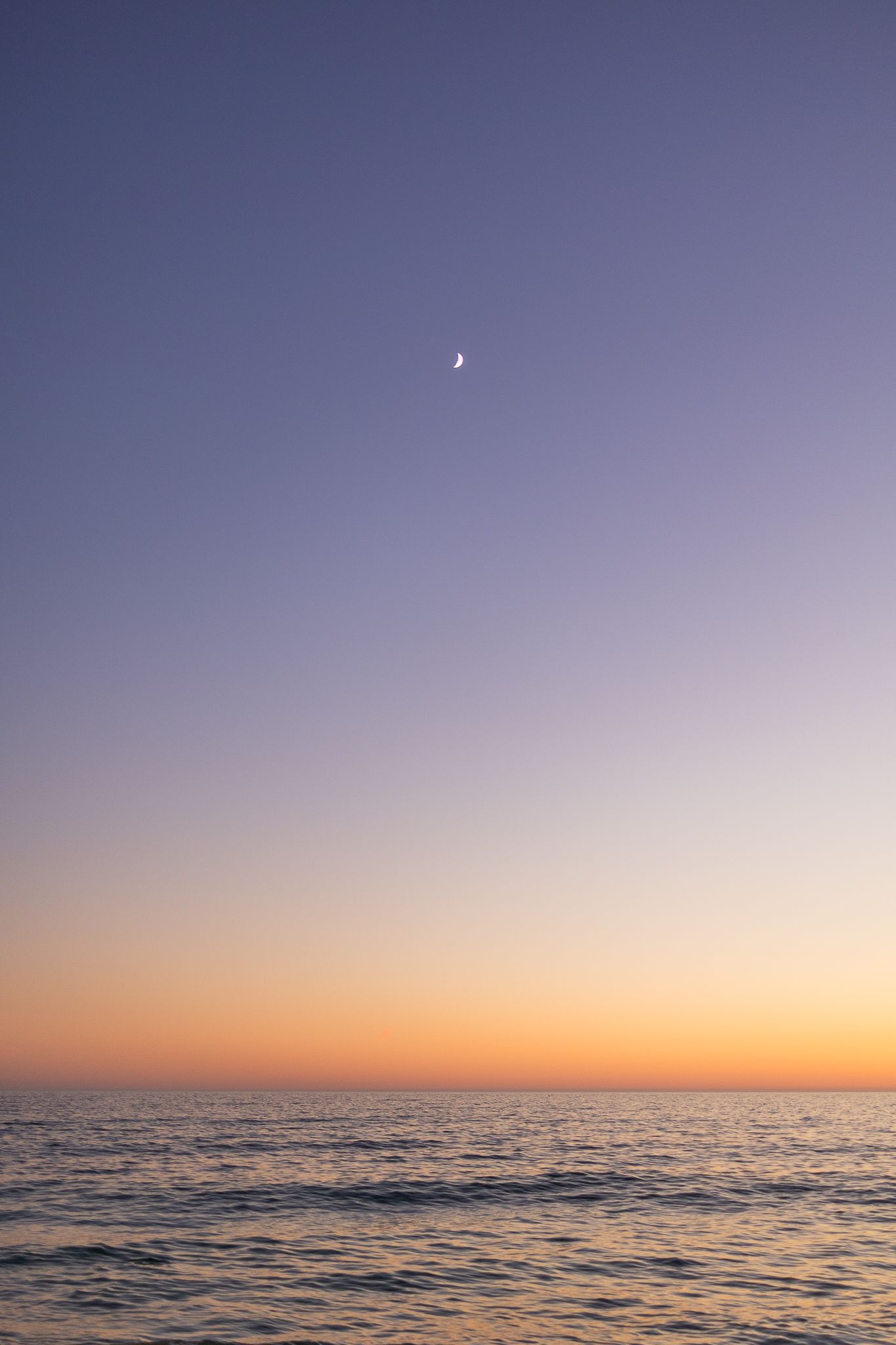 Tranquil ocean sunset with a crescent moon visible in the purple and orange gradient sky.