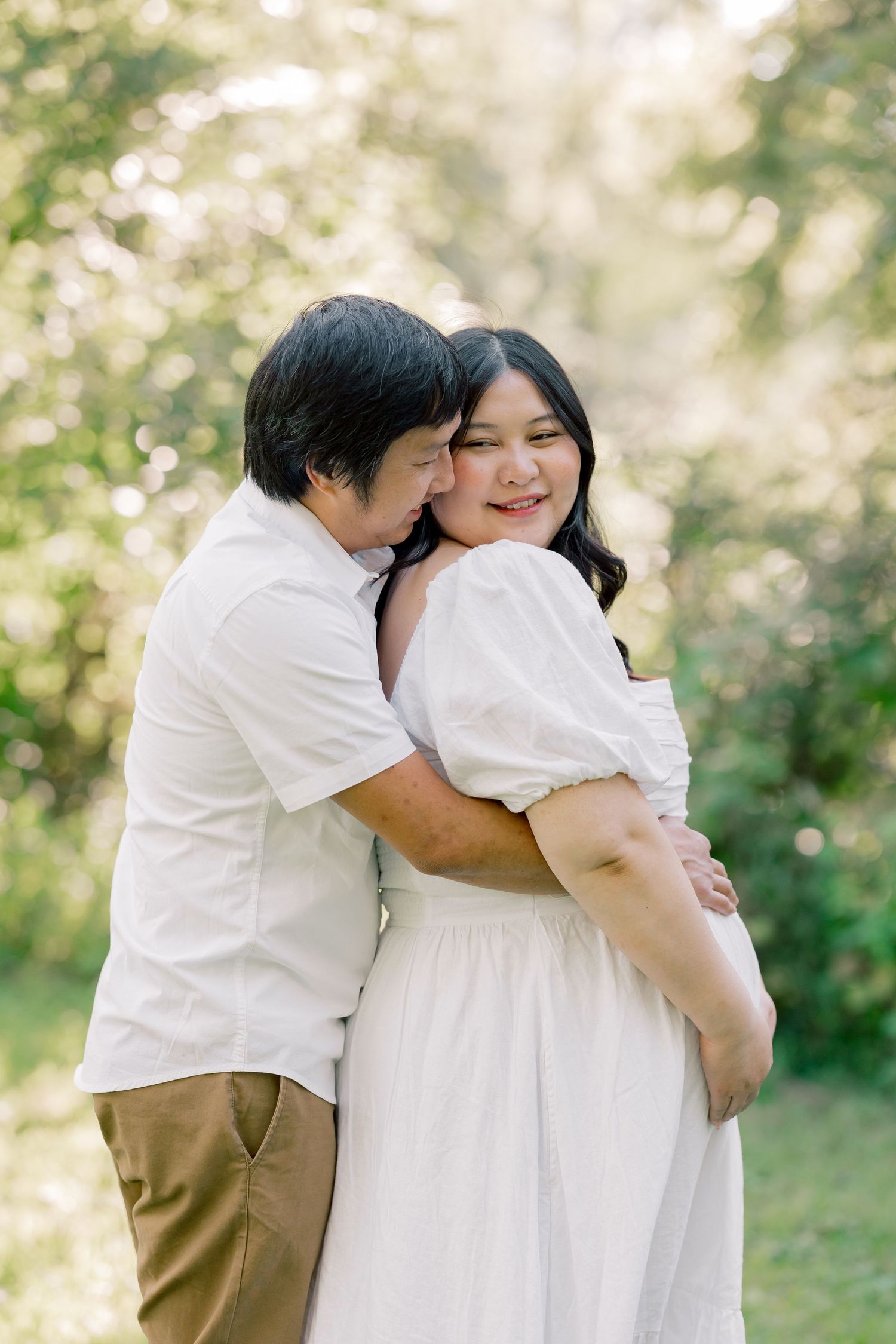 A tender embrace between a loving couple wearing white outfits in a sun-dappled outdoor setting.