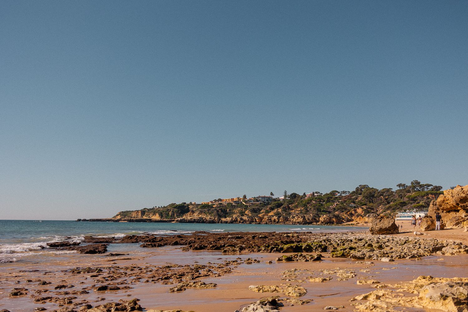 Golden sandy beach with rocky outcrops and coastal cliffs under clear blue skies along the Algarve coastline.