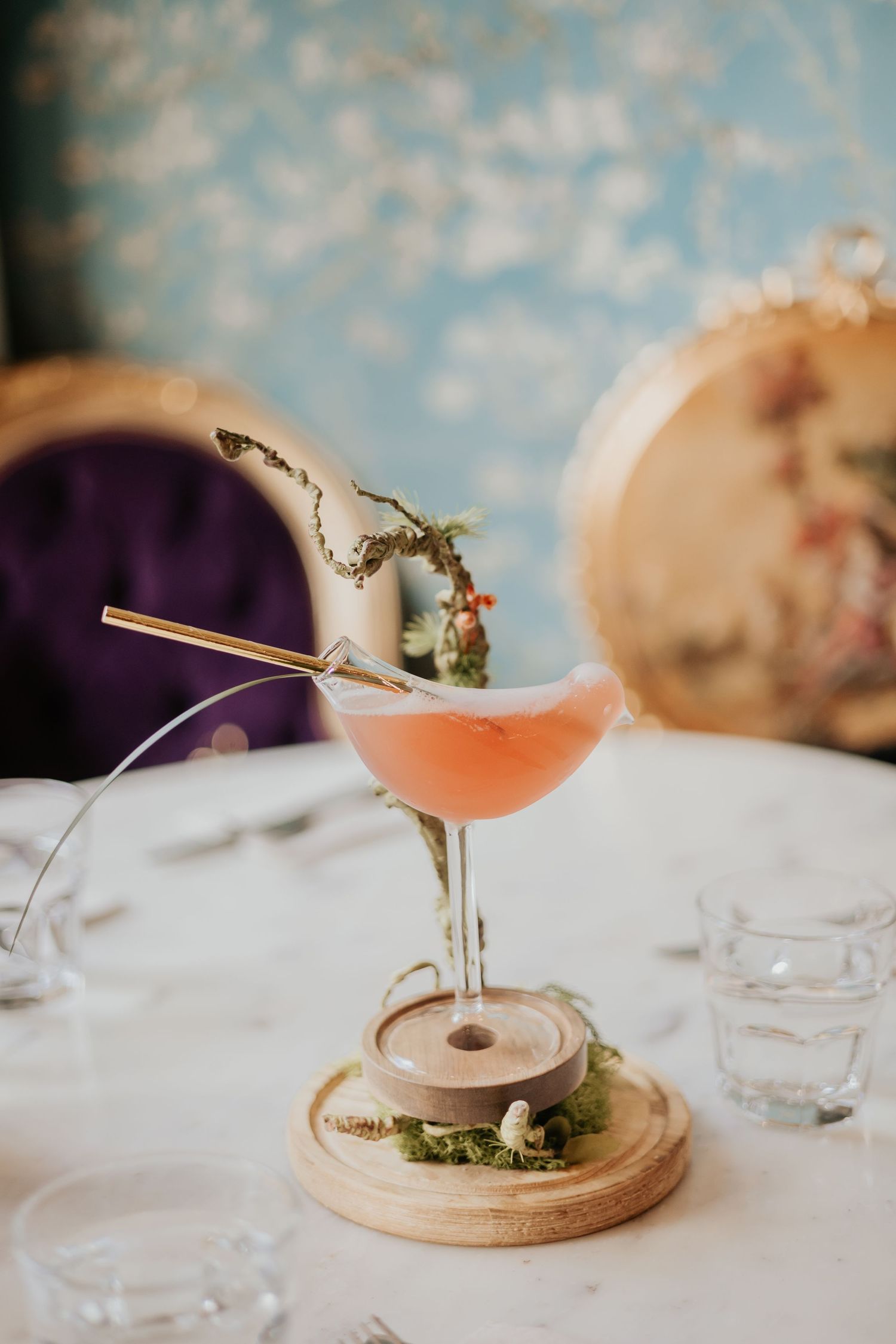 Pink cocktail served in a coupe glass with botanical garnish on wooden coaster against blurred background.