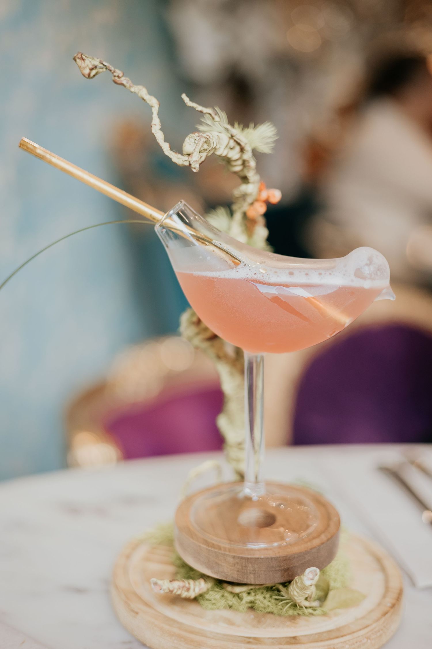 A pink cocktail served in a coupe glass on a wooden stand with dried floral garnish.