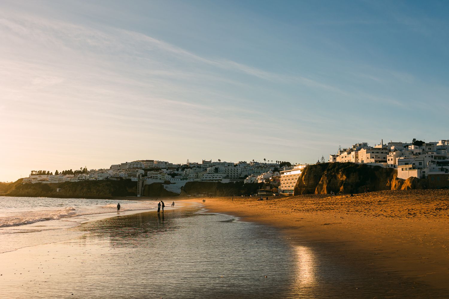 Golden sandy beach at sunset with cliffside houses overlooking the shoreline and people walking.