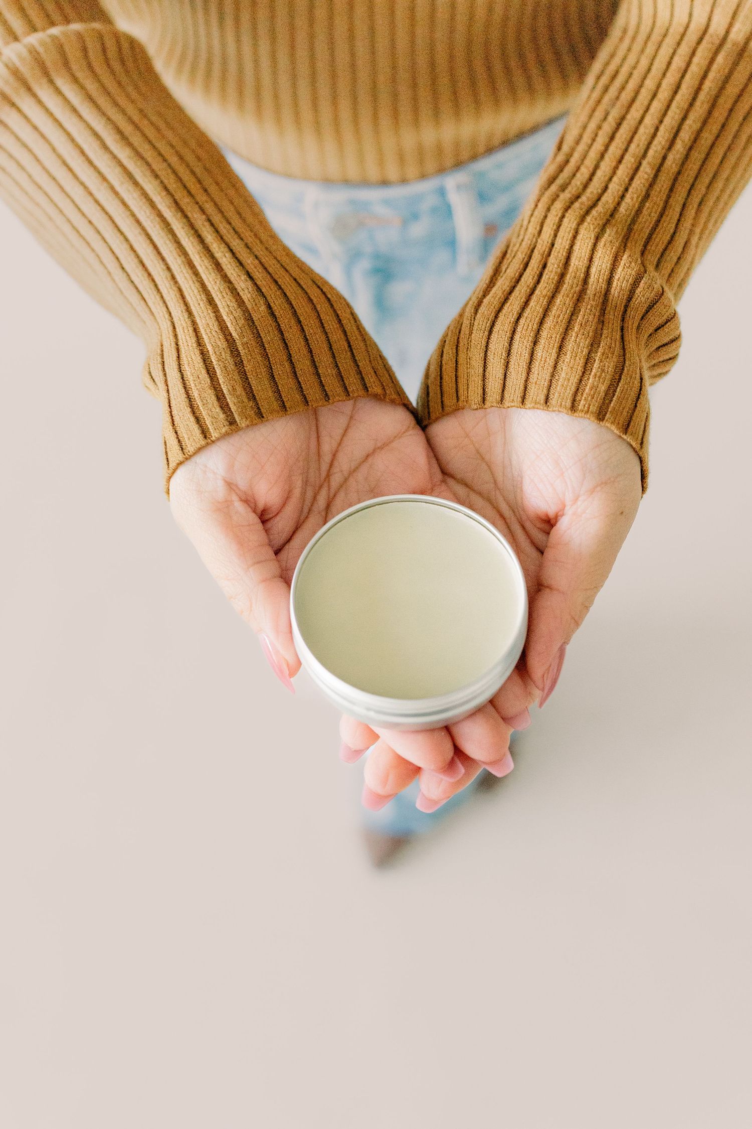 Hands wearing brown ribbed sweater sleeves hold a small white ceramic cup or bowl against a light background.