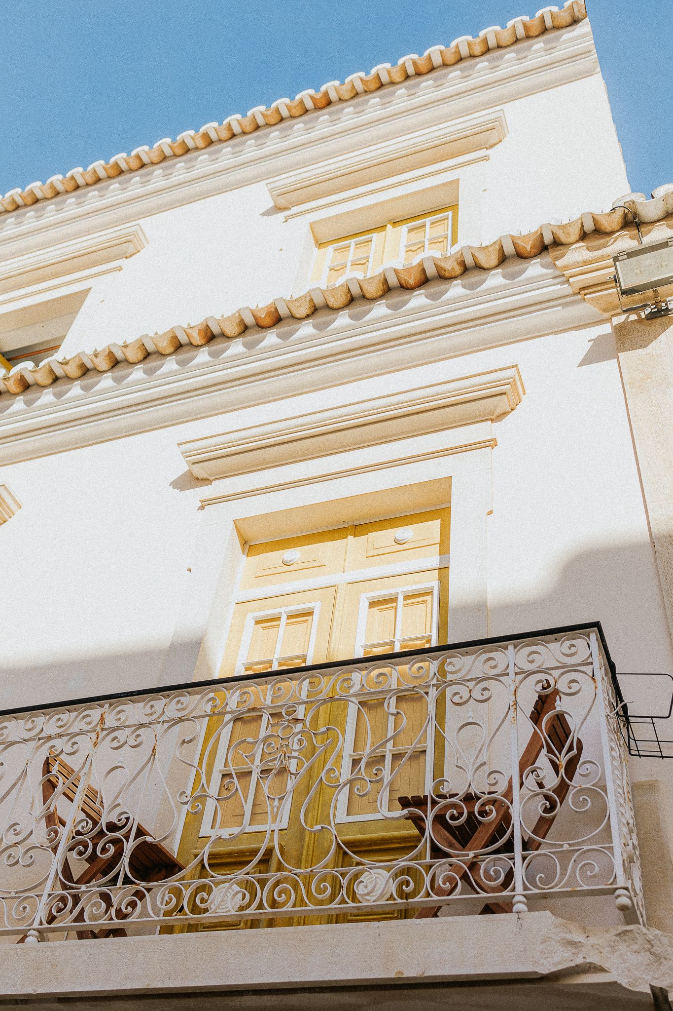 A traditional Mediterranean building with white walls, ornate wrought iron balcony, and decorative yellow trim against blue sky.