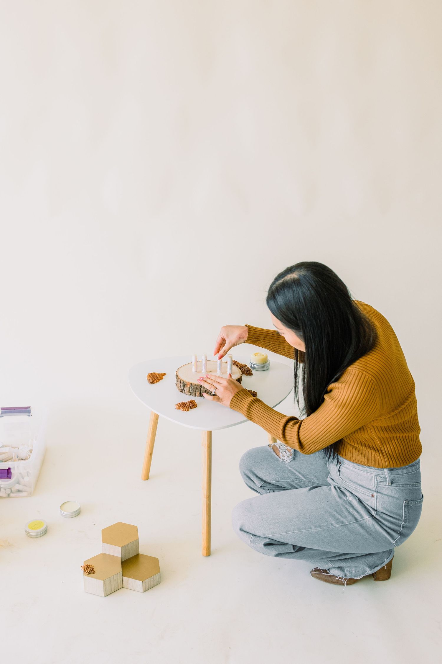 Person in mustard sweater and jeans crouches to photograph wooden blocks on small white table against neutral wall.