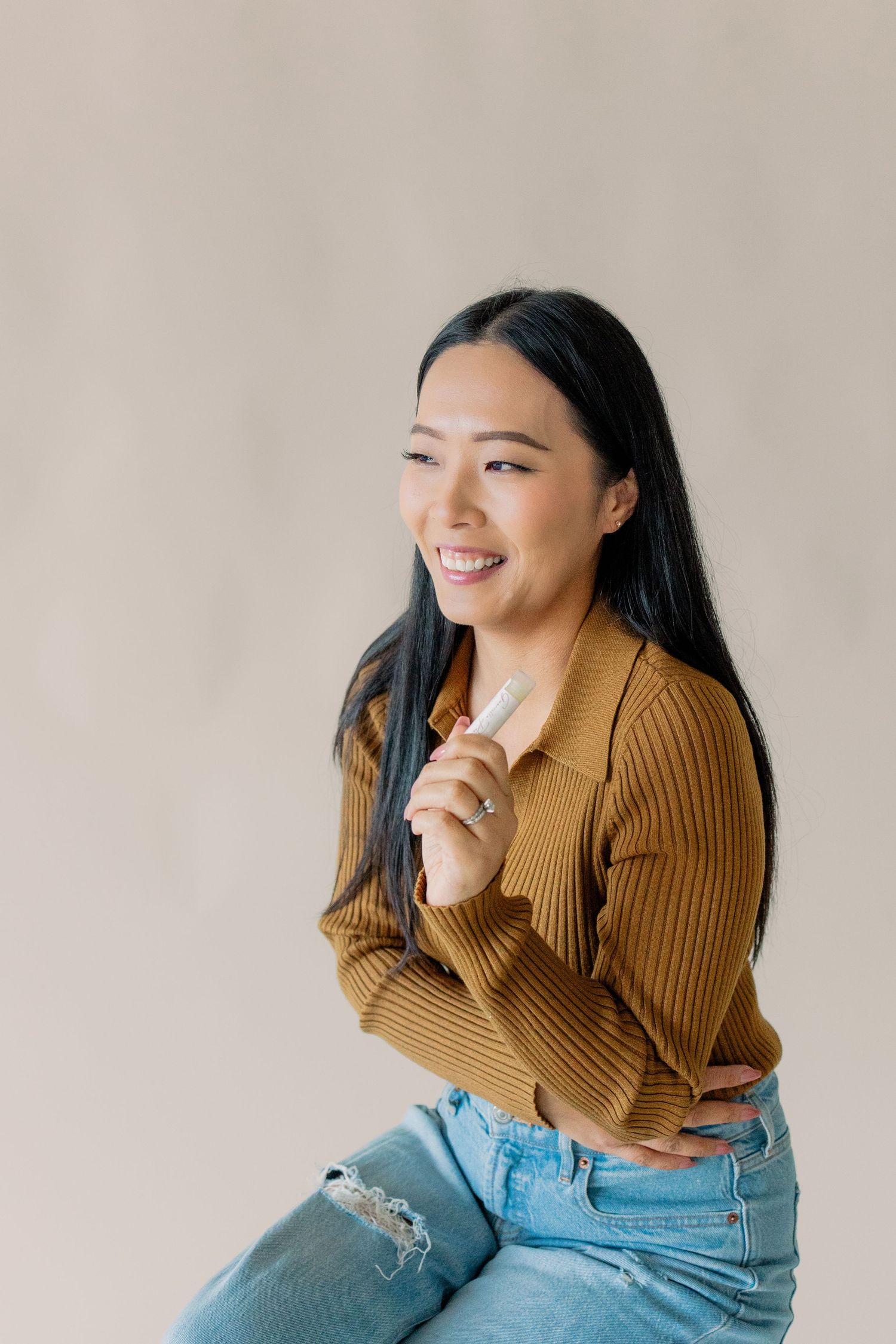 Woman in tan ribbed sweater and ripped jeans poses against light background while smiling and laughing.
